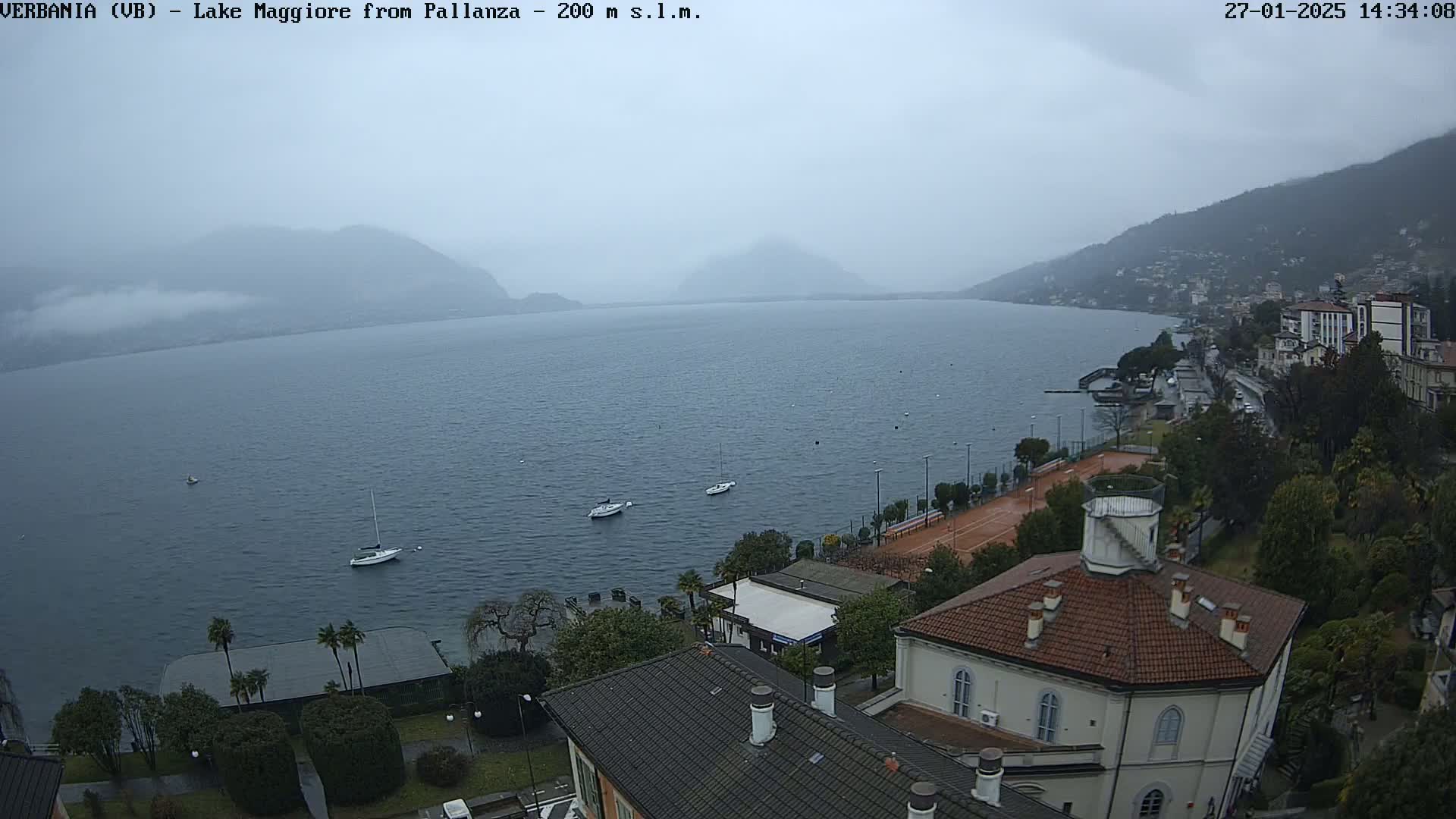 A hazy, overcast view of Lake Maggiore from Pallanza shows several small boats on the water, a tennis court, and buildings along the shoreline.