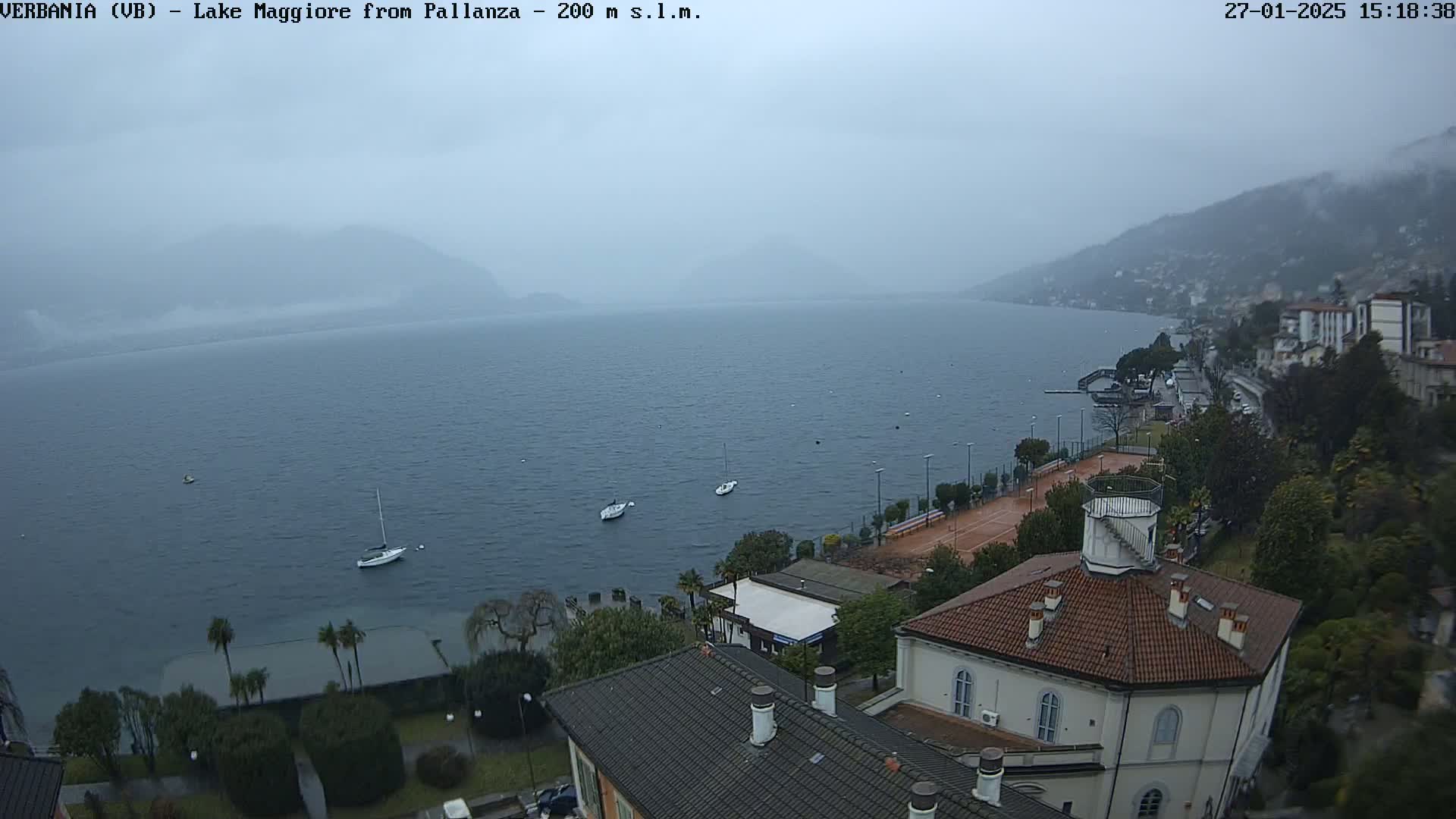 A hazy, overcast day reveals a lake with a few sailboats, a shoreline with buildings and trees, and mountains in the distance.