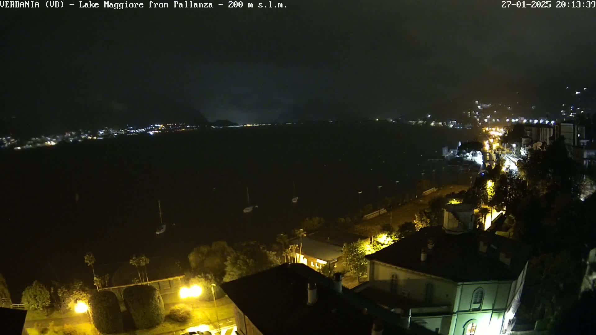 A nighttime view of a lake with a few boats and a town with lights illuminated along the shoreline under a dark, cloudy sky.