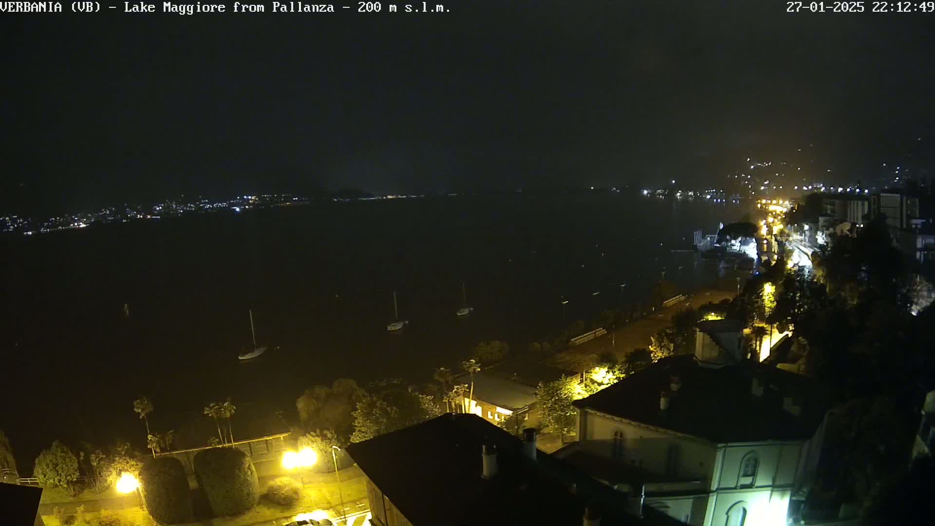 A nighttime view of a dark lake with a few boats, a shoreline with buildings and streetlights, and a distant city skyline under a cloudy sky.