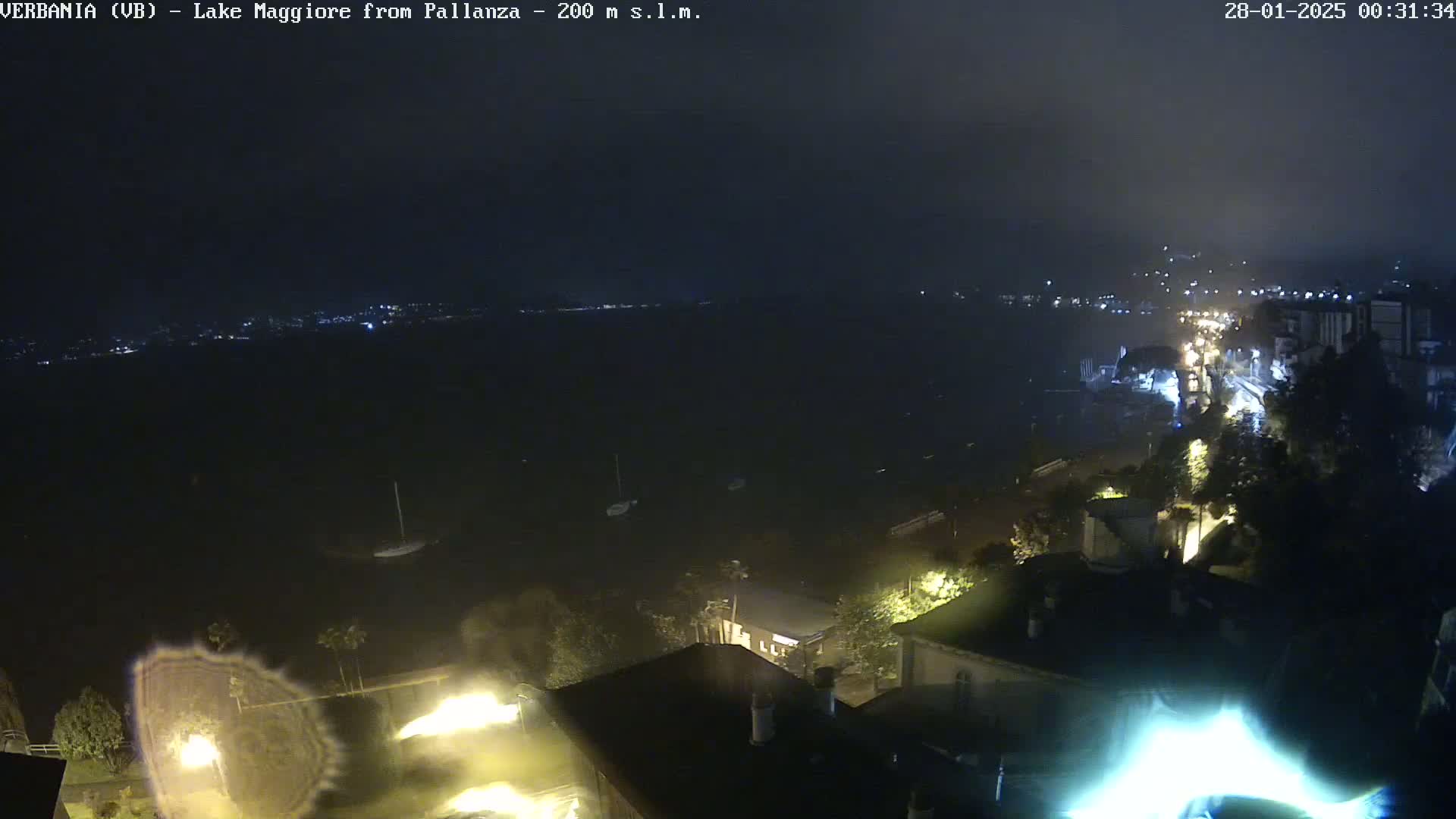 A nighttime, high-angle view shows a dark body of water with a town's lights visible across it and along a shoreline, under a mostly cloudy sky.