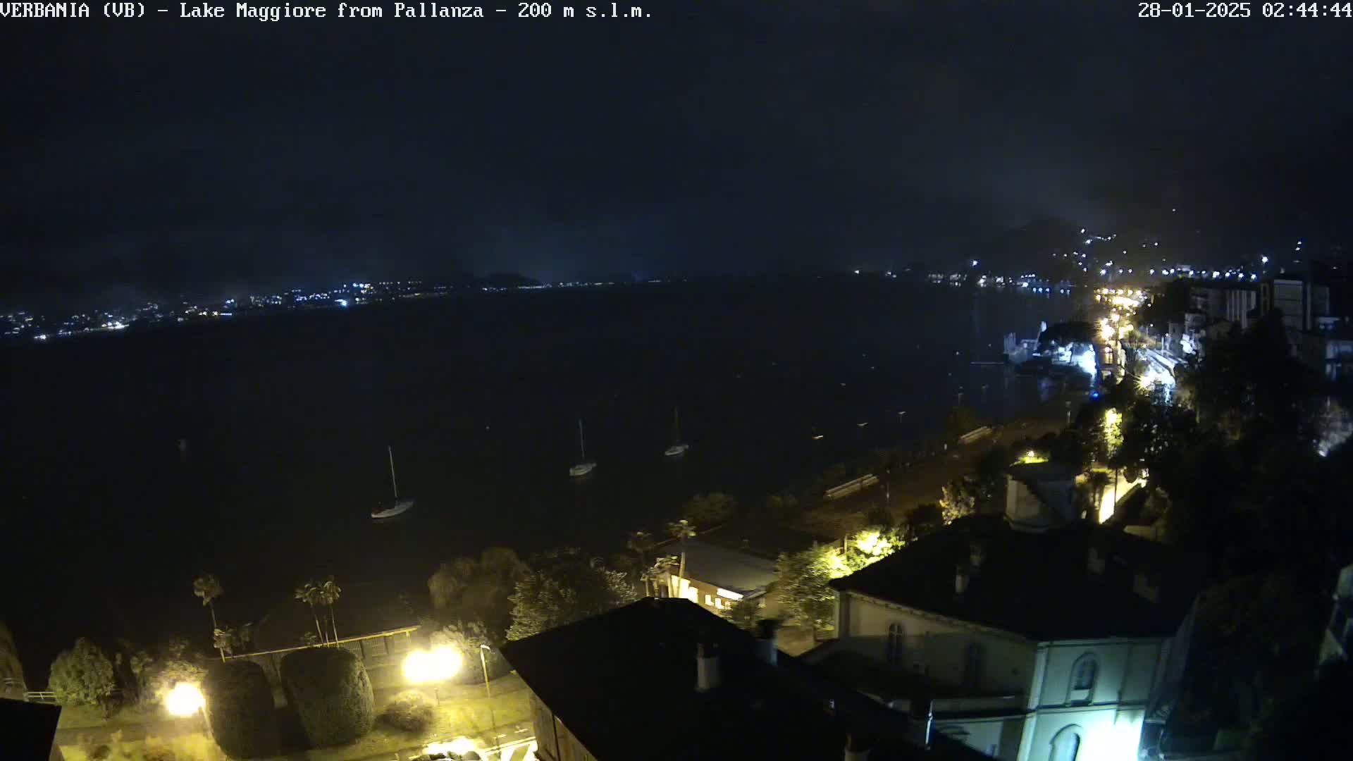 A nighttime view of a lake with a few boats, a shoreline with buildings and lights, and a dark, cloudy sky.