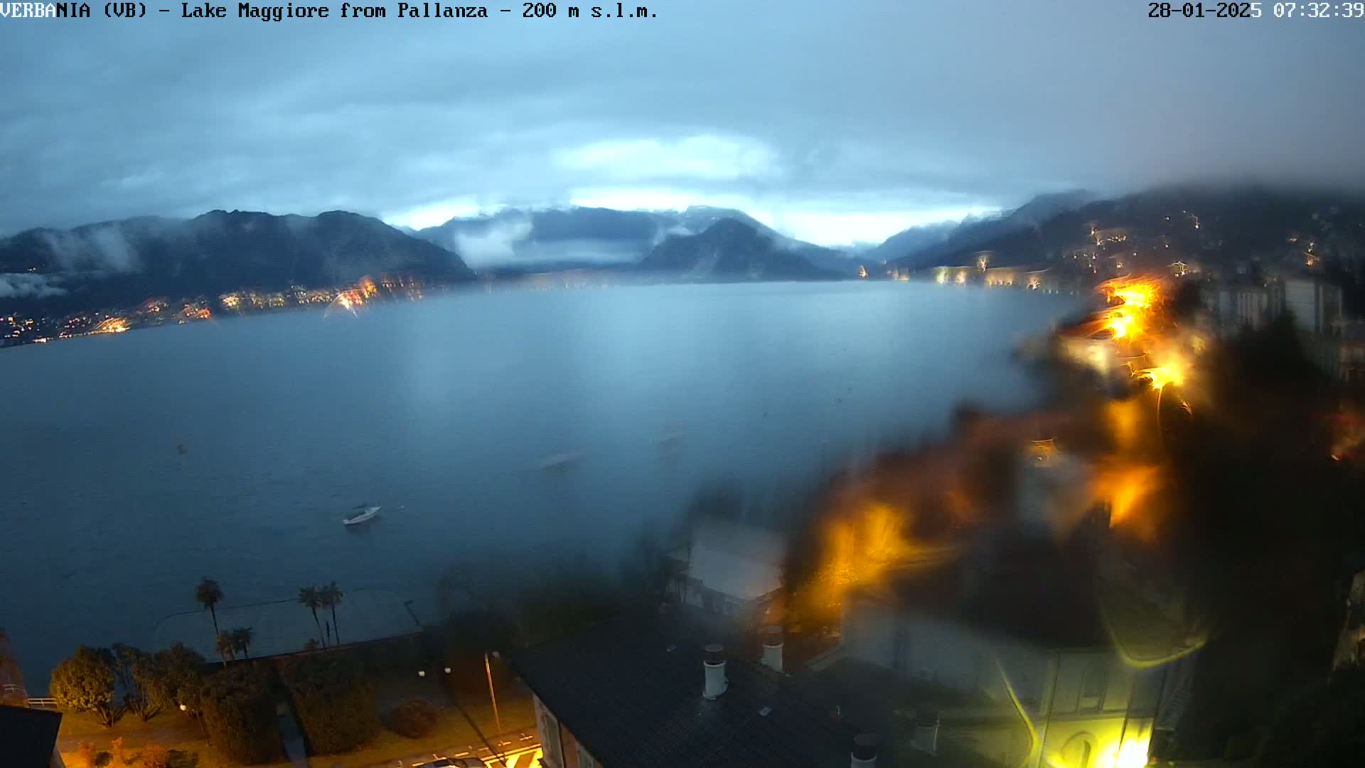 A rain-streaked window reveals a view of Lake Maggiore at twilight, with mountains shrouded in mist in the background and lights illuminating a lakeside town.