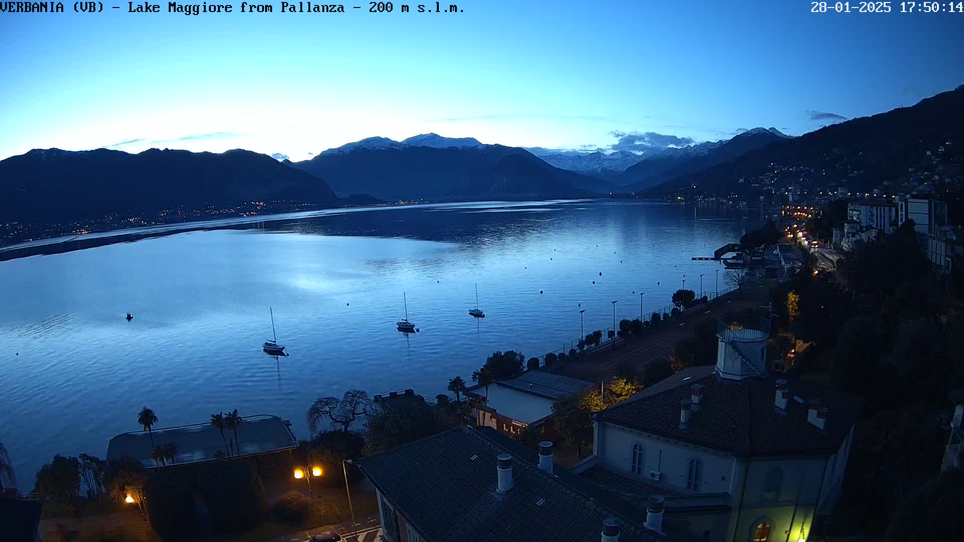 A twilight view of a calm lake with snow-capped mountains in the background, a few sailboats on the water, and a lakeside town with lights illuminated at dusk.