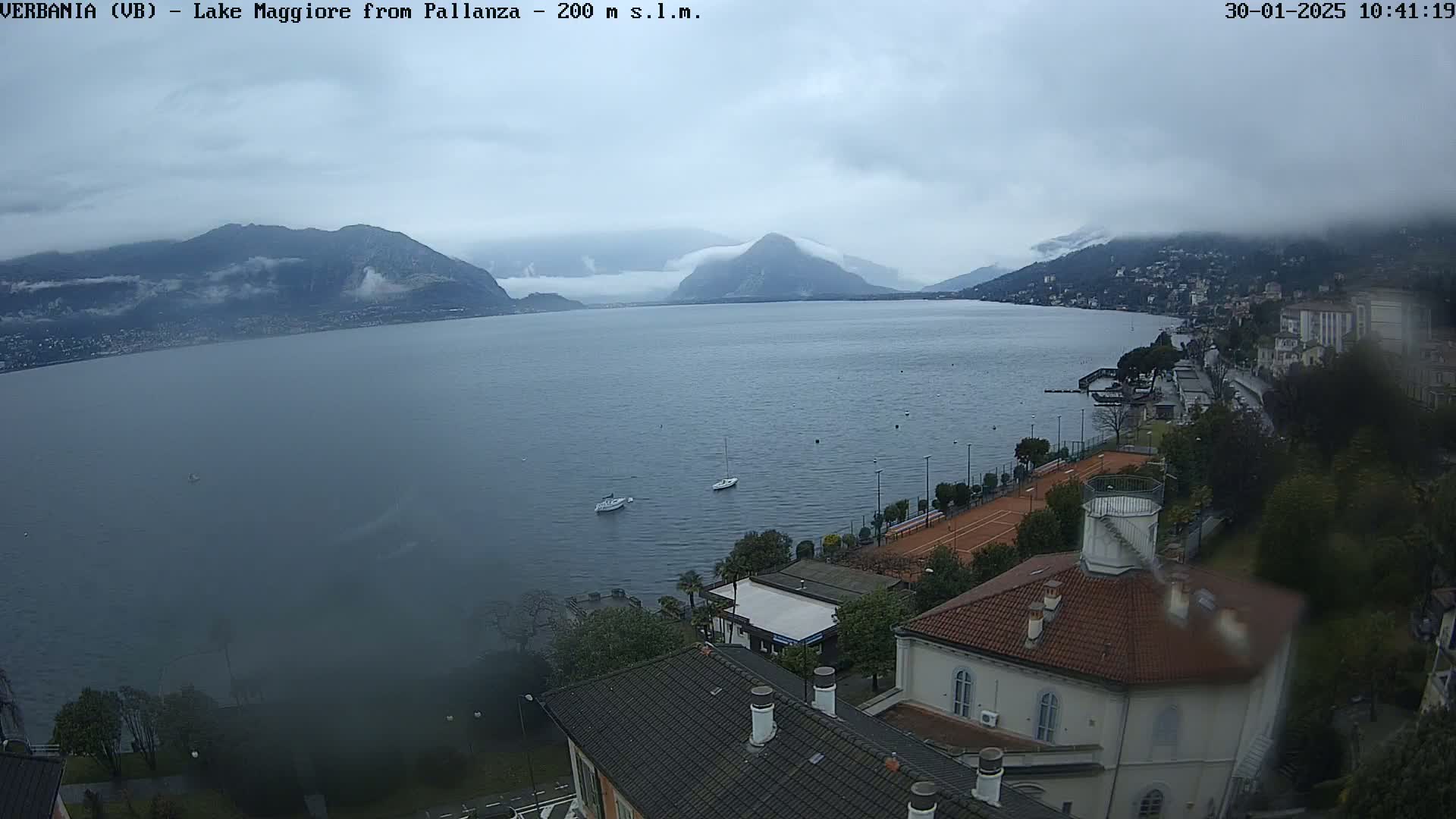 A cloudy, overcast day reveals a view of Lake Maggiore from a high vantage point, showing a shoreline with buildings, tennis courts, and a few boats on the water.