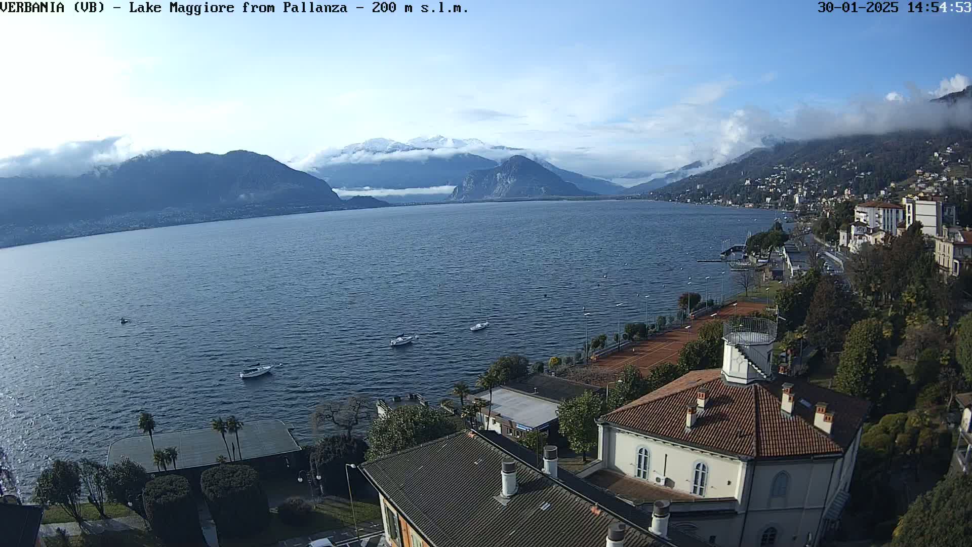 A partly cloudy day overlooks a large lake with snow-capped mountains in the distance, a few boats on the water, and buildings and tennis courts along the shoreline.