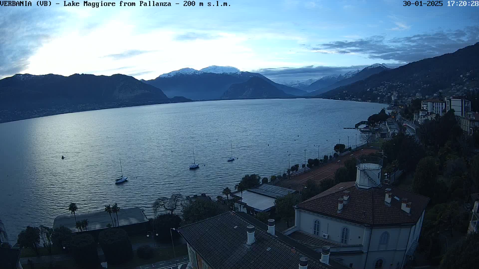 A calm lake at dusk, surrounded by snow-capped mountains and a lakeside town, with a few sailboats visible on the water.