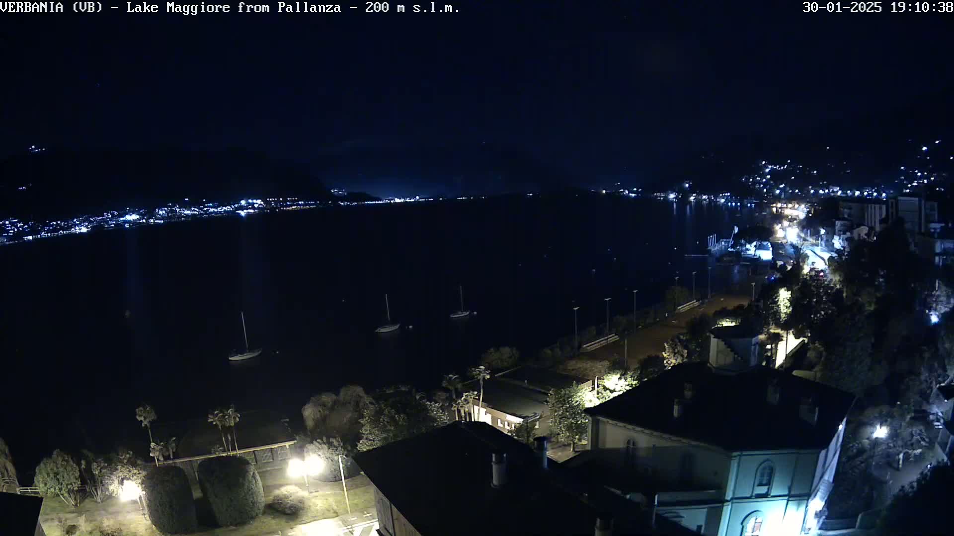 A nighttime, aerial view of a dark lake with a few boats, illuminated buildings along the shoreline, and hills in the distance under a clear sky.