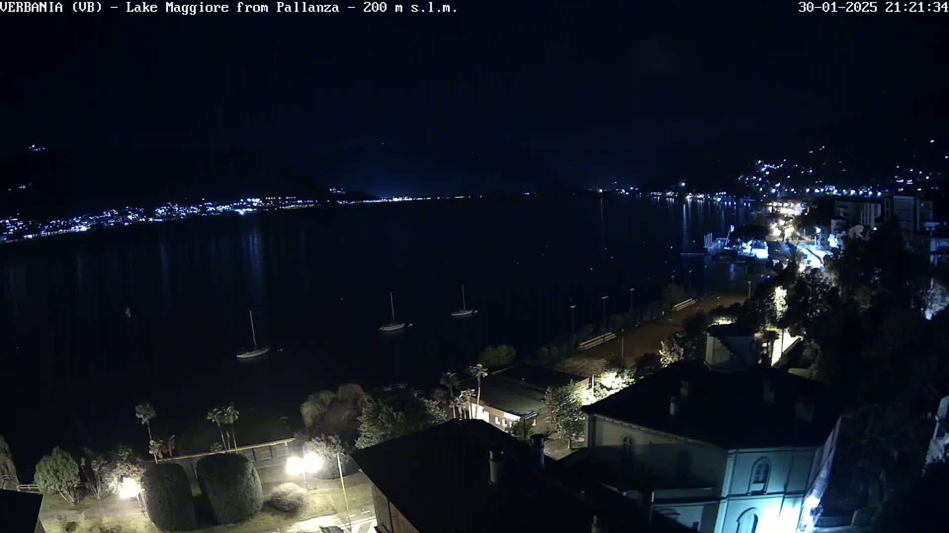 A nighttime view of a lake with a town's lights reflecting on the water, several boats moored, and buildings illuminated along the shore.