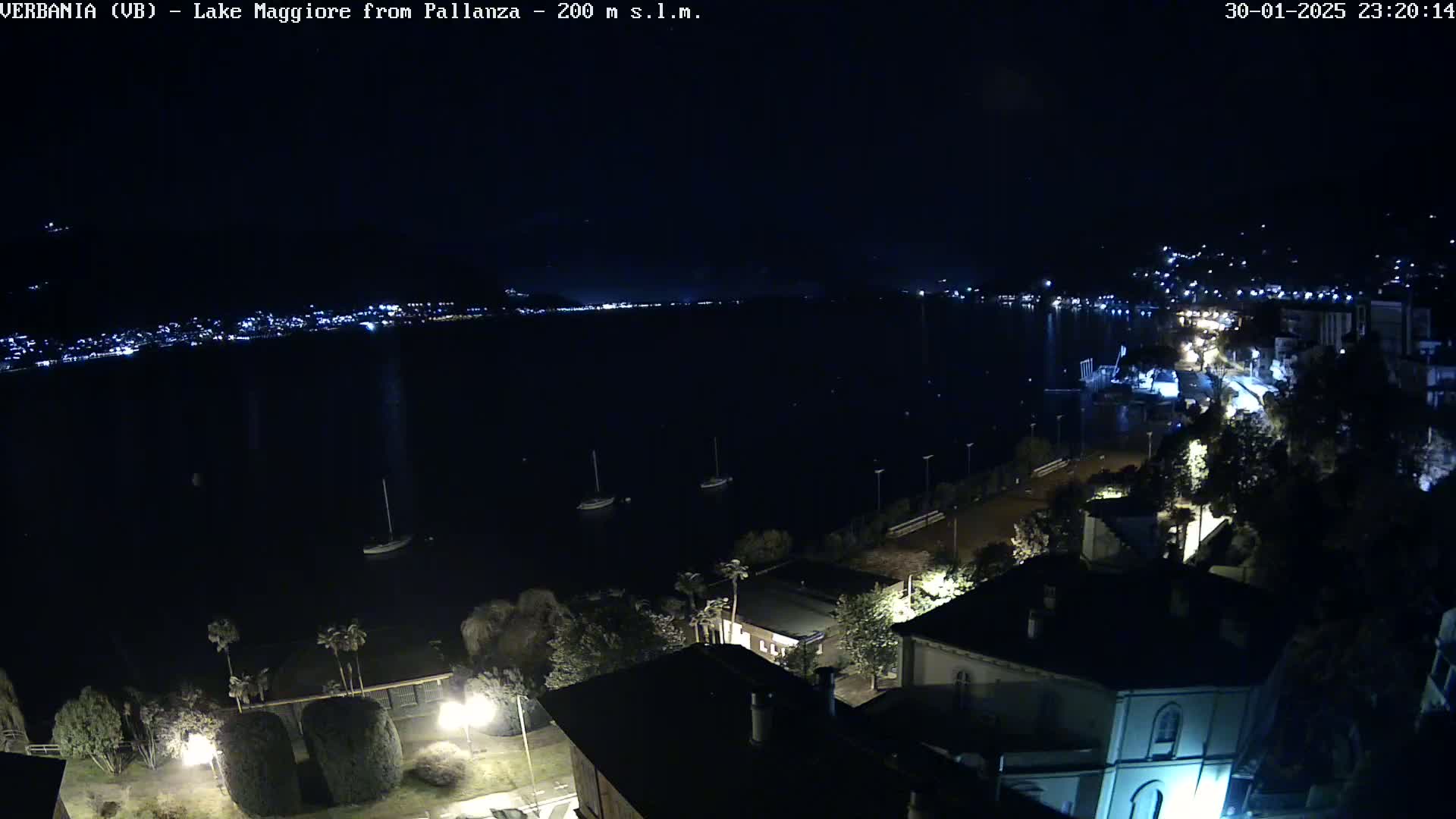 A nighttime view of a lake with city lights across the water and a few boats, from a vantage point overlooking buildings and trees.