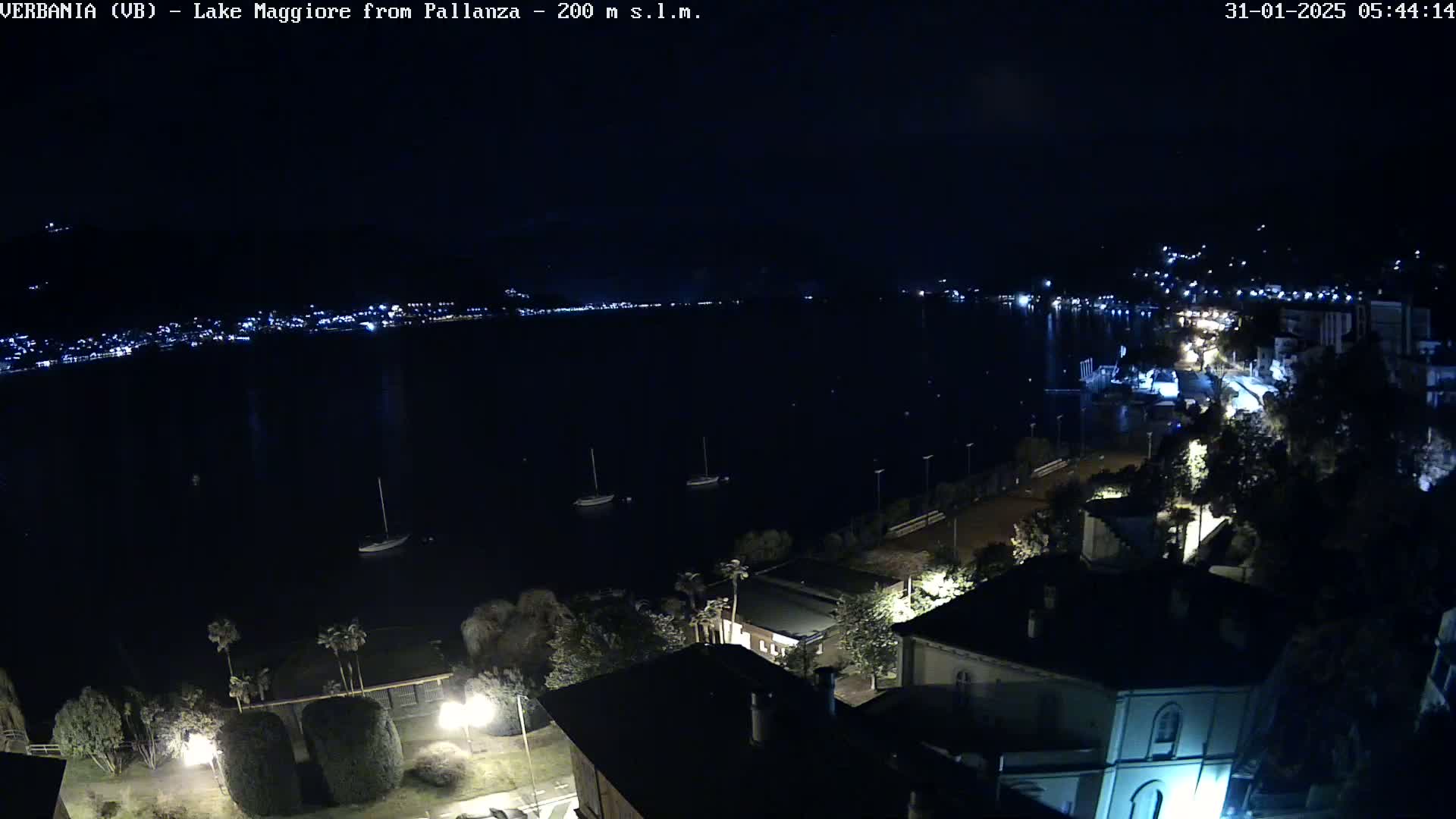 A nighttime, high-angle view of a lake with a town's lights visible across the water, several boats, and buildings and trees in the foreground.