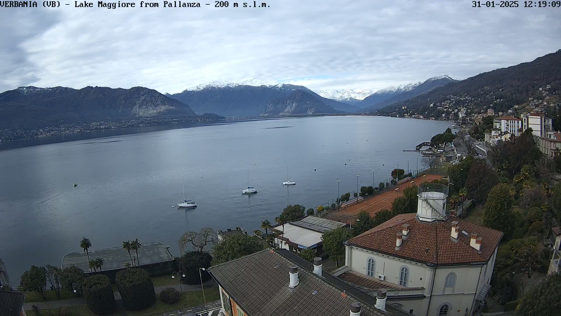 A partly cloudy day reveals a lake with several sailboats, bordered by buildings and tennis courts, with snow-capped mountains in the background.