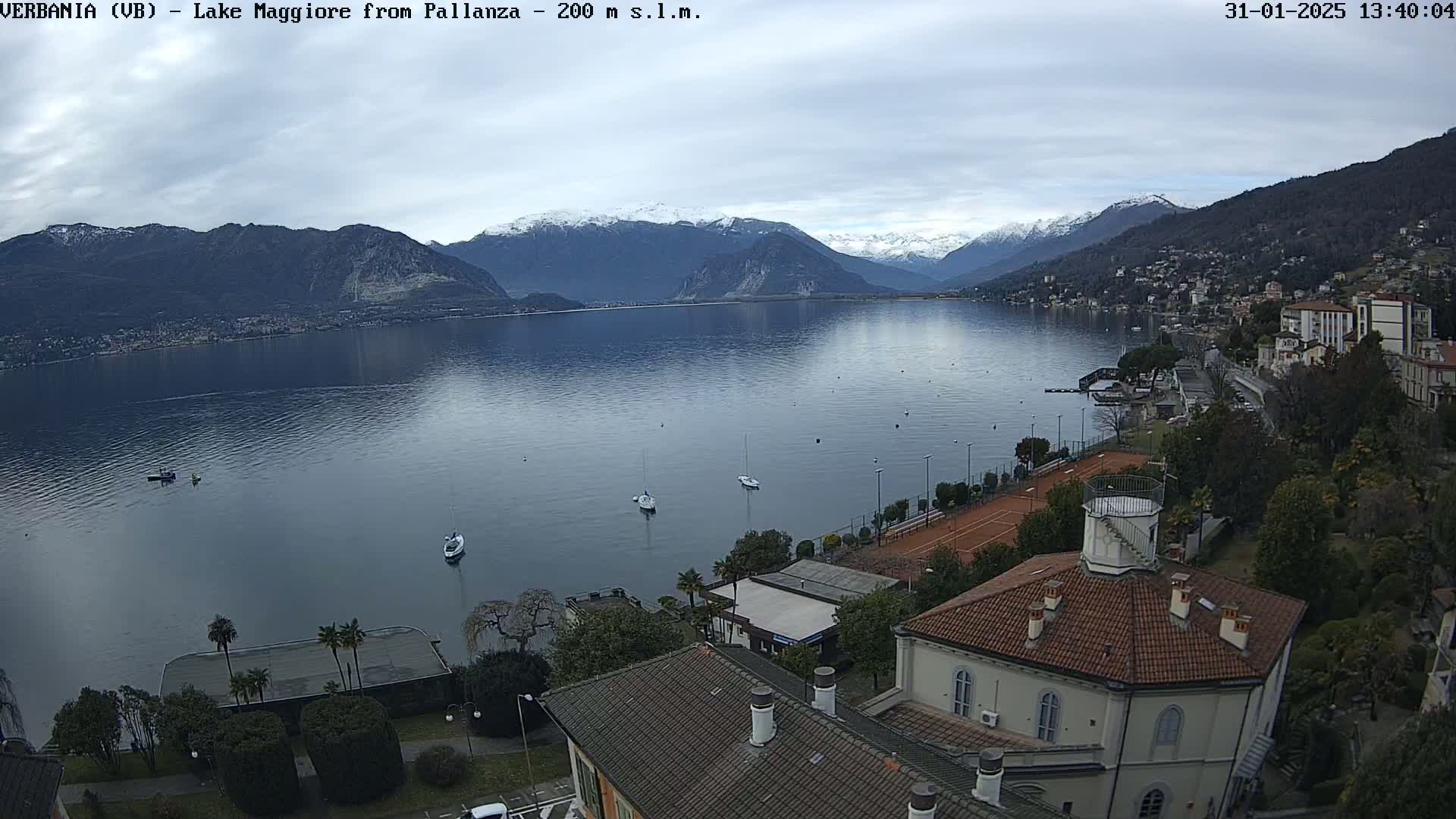 A calm lake stretches towards snow-capped mountains under an overcast sky, with a shoreline featuring buildings, tennis courts, and several small boats.