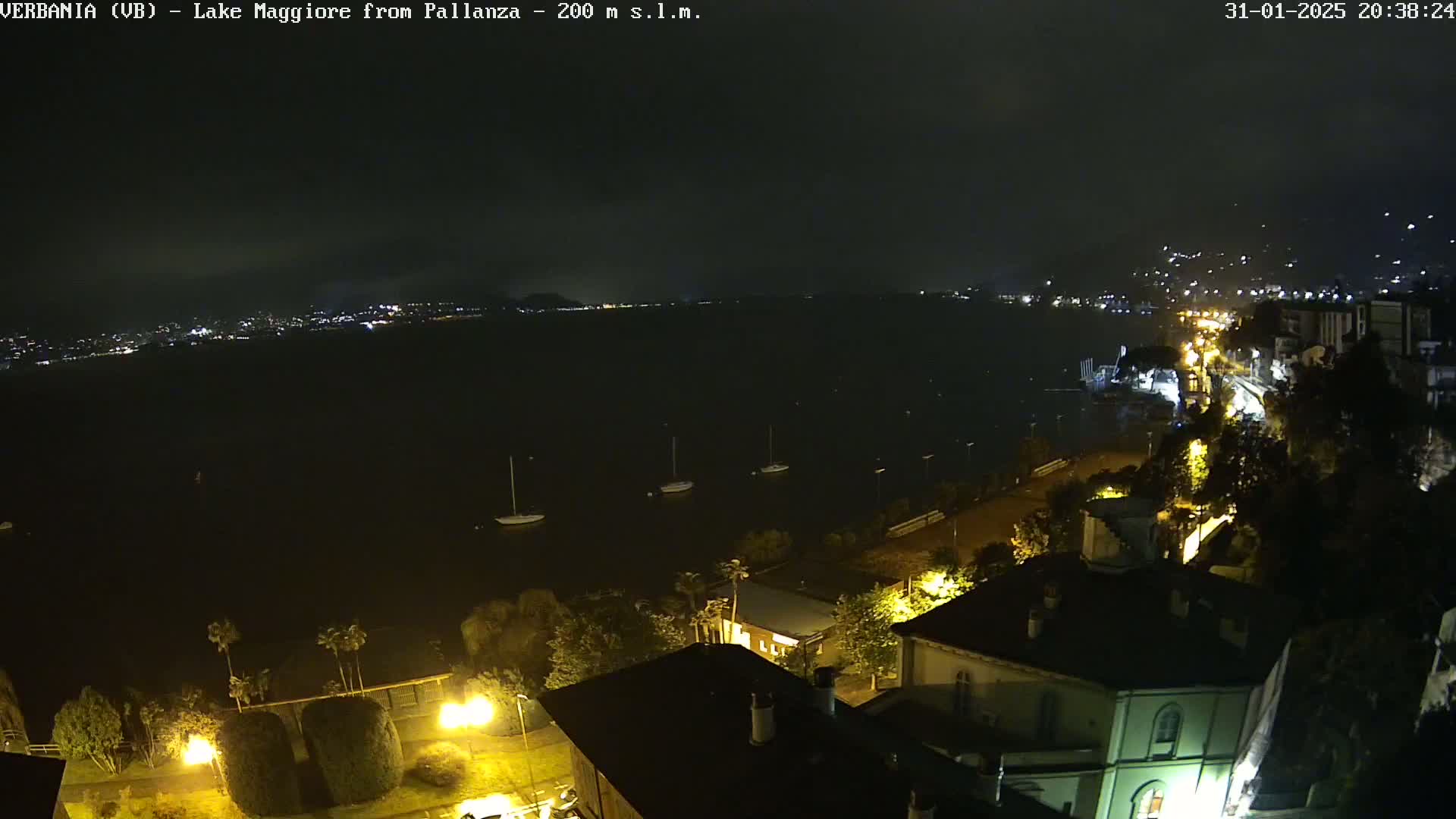 A nighttime view of a lake with several sailboats, a town's lights illuminating the shoreline, and dark clouds overhead.