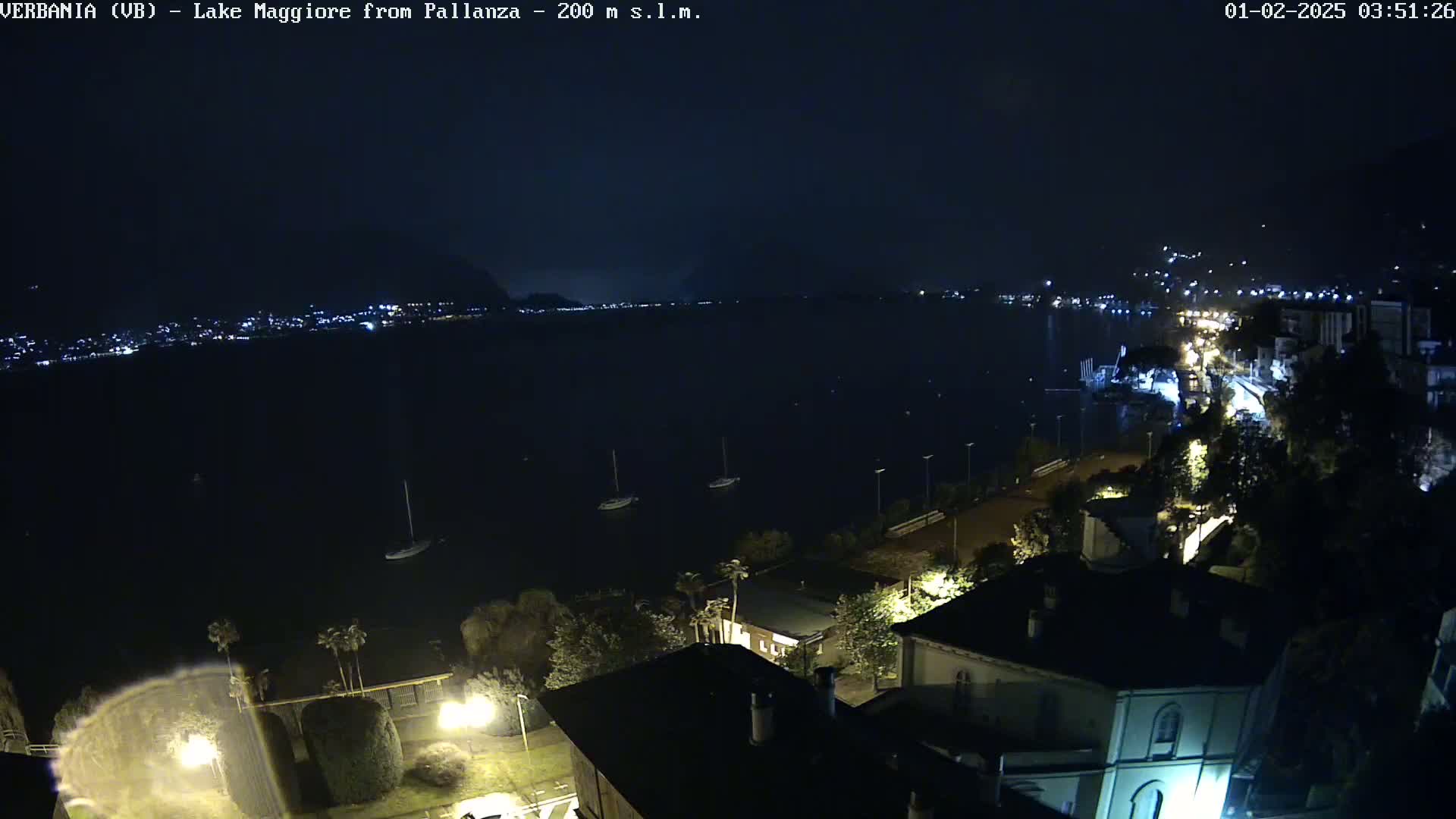 A nighttime view of a calm lake with a few boats, illuminated buildings along the shoreline, and dark mountains in the distance.