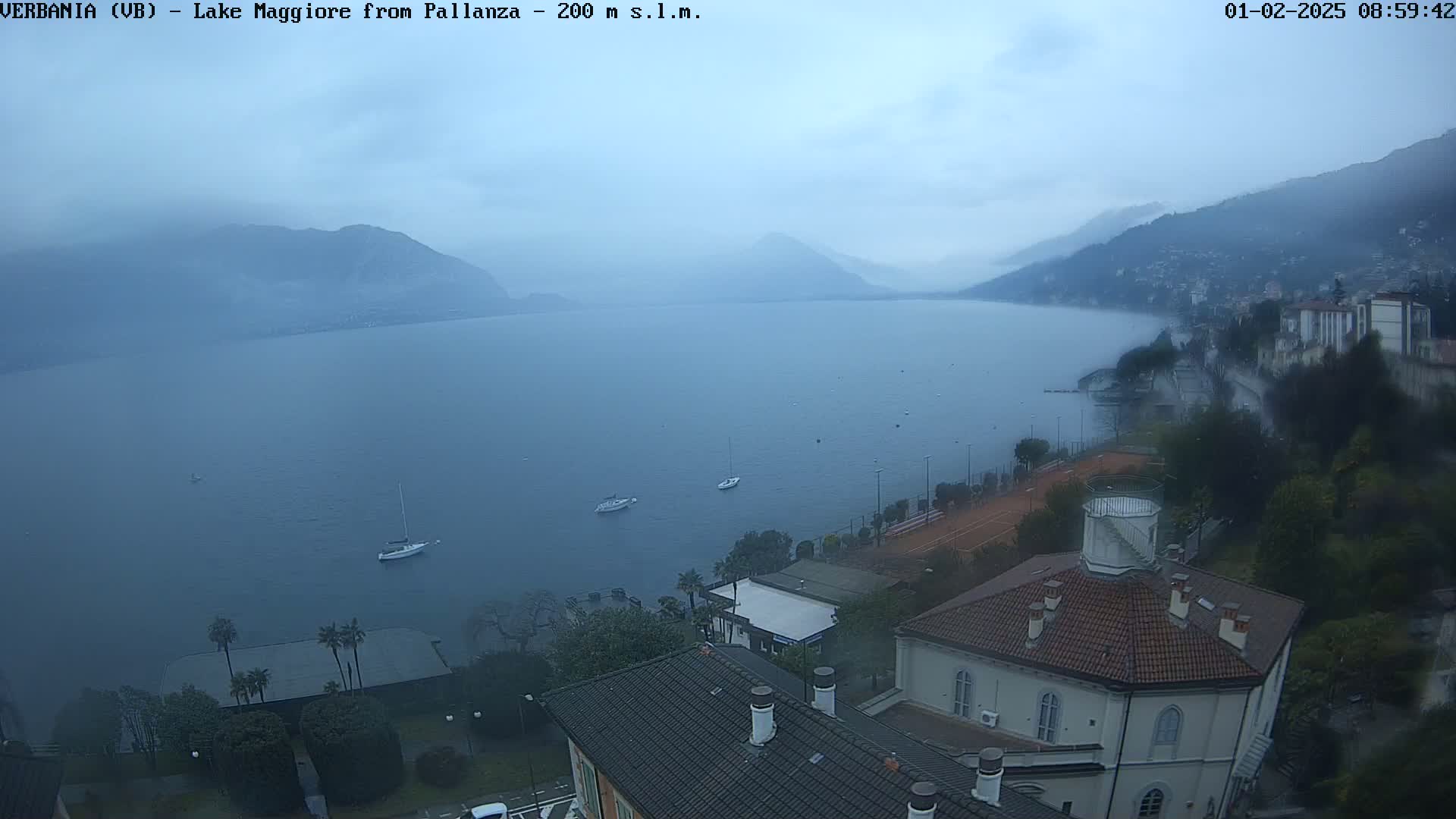 A hazy, overcast day reveals a view of Lake Maggiore from Pallanza, with several boats on the water and buildings along the shoreline.