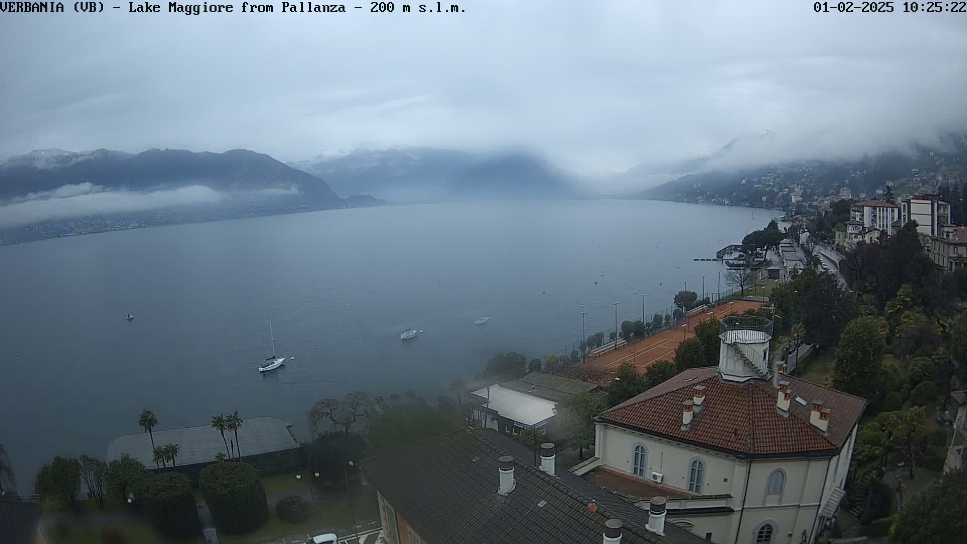 A hazy, overcast day reveals a lake surrounded by mountains partially obscured by fog, with a few boats on the water and buildings along the shoreline.