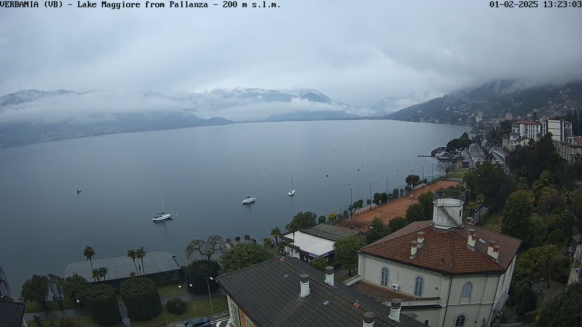 An overcast day reveals a lake, dotted with sailboats, bordered by buildings and tennis courts, with snow-capped mountains in the background.