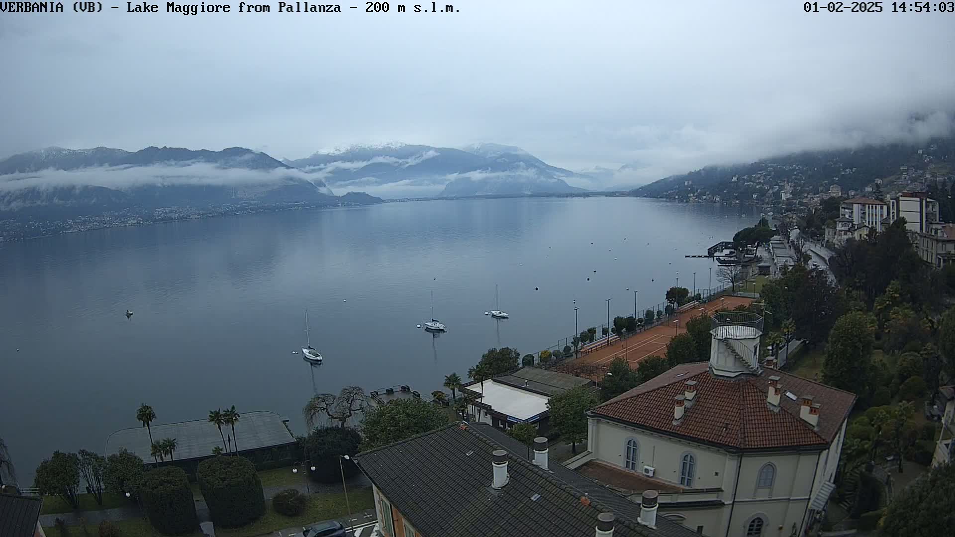 A lake, partially shrouded in fog, is seen from an elevated vantage point, with several boats on the water, buildings along the shore, and mountains in the background under an overcast sky.