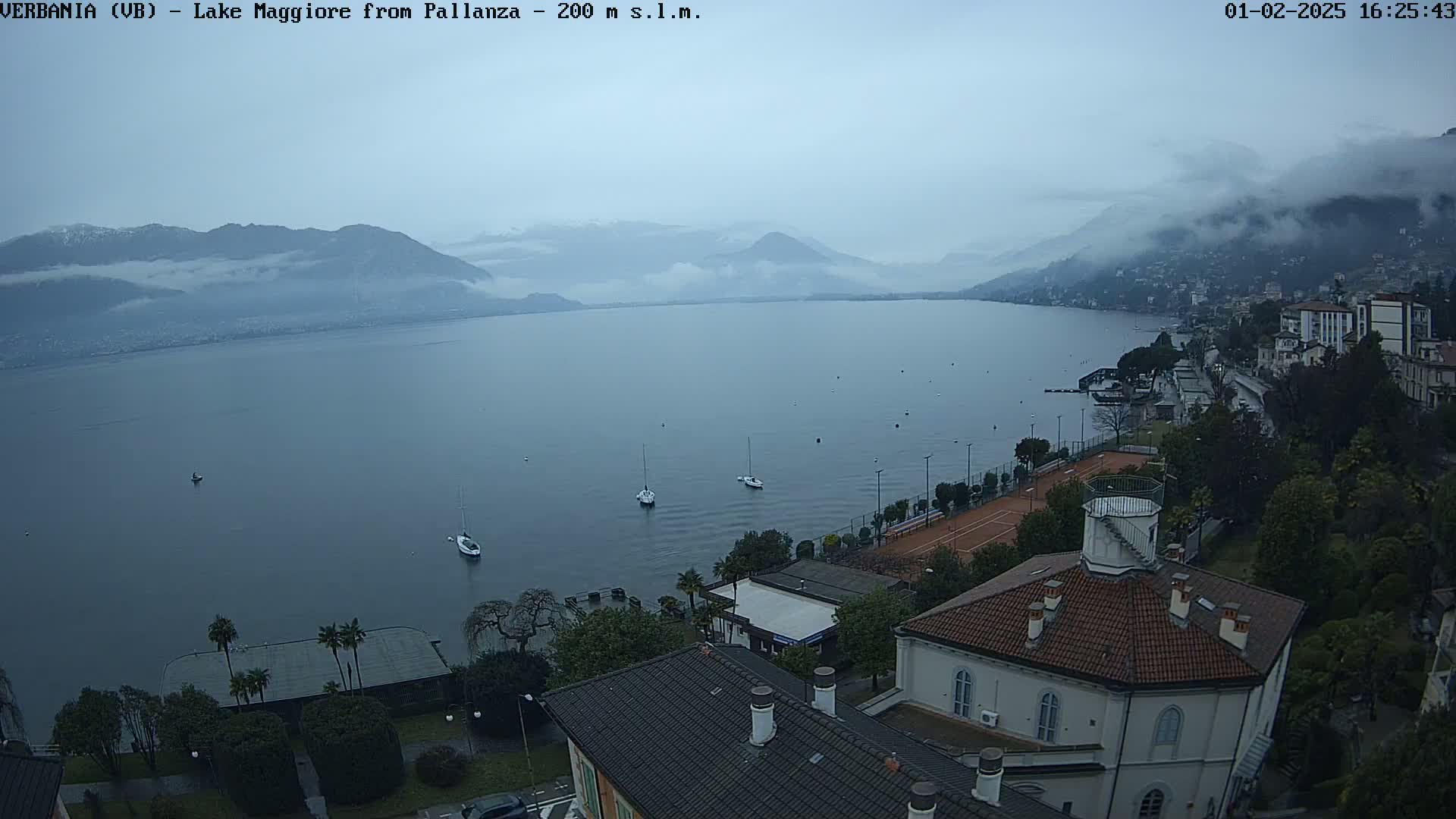 A large lake is seen from an elevated position on an overcast day, with several sailboats visible on the water, a shoreline with buildings and trees, and mountains shrouded in fog in the background.