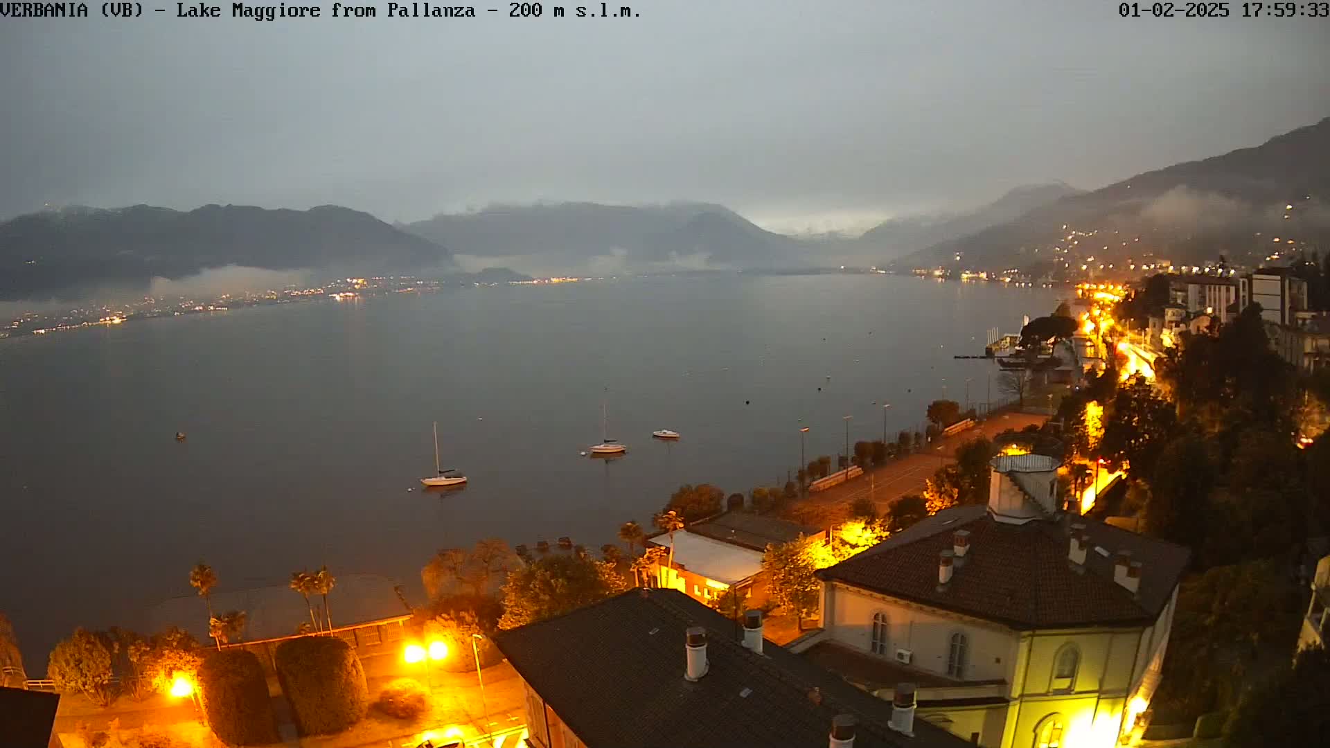 A nighttime, hazy view of Lake Maggiore from Pallanza shows a few sailboats on the water, illuminated buildings along the shore, and mountains shrouded in mist in the background.