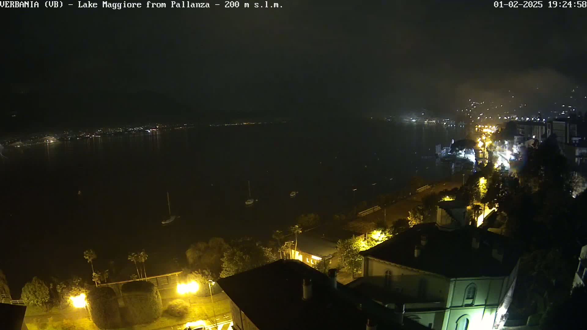 A nighttime view of a lake with a town's lights illuminating the shoreline, a few boats on the dark water, and buildings with illuminated windows visible along the slope.