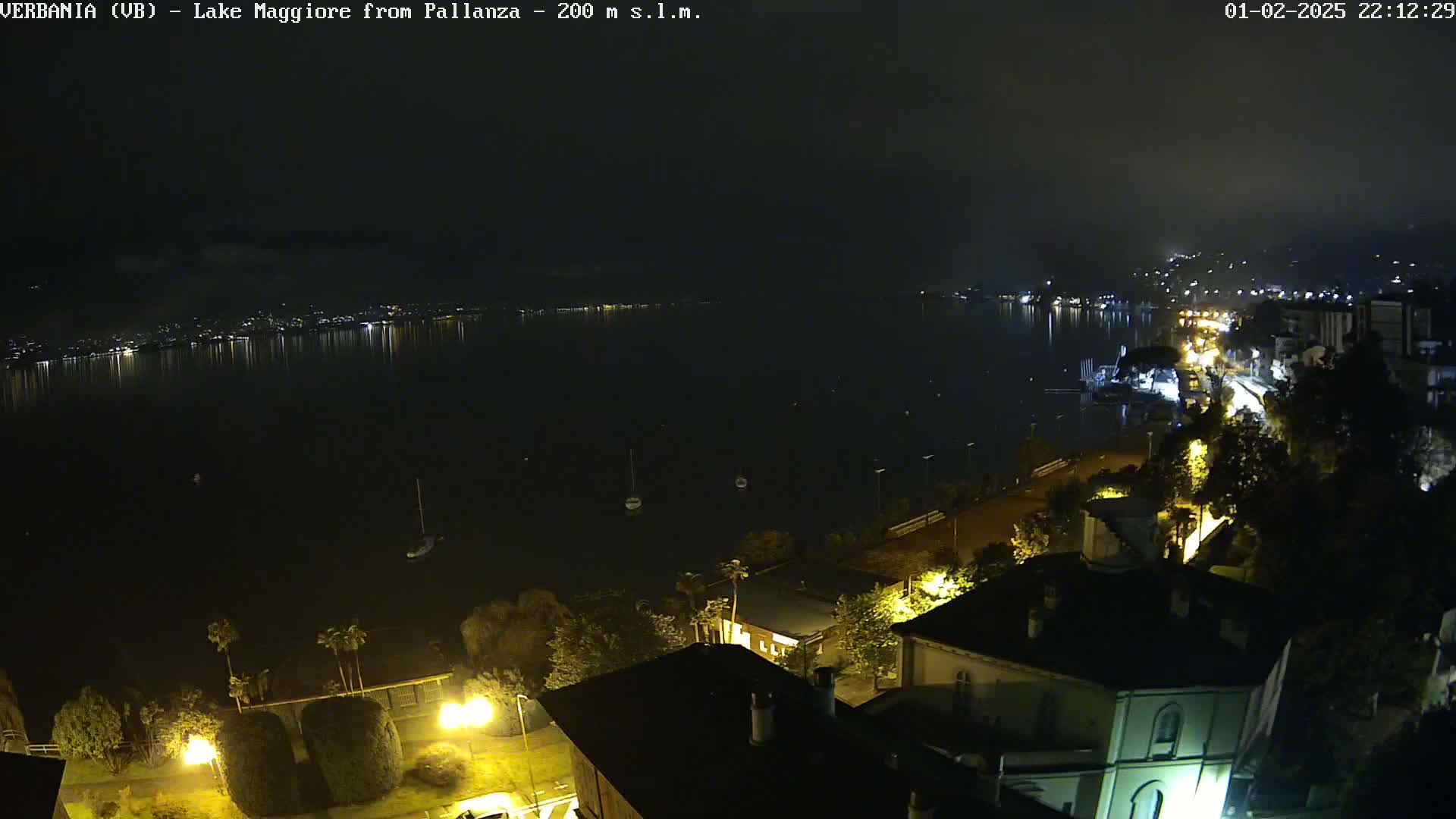 A nighttime, slightly hazy view of a lake with lights from a town across the water and buildings and streetlights along the shore in the foreground.