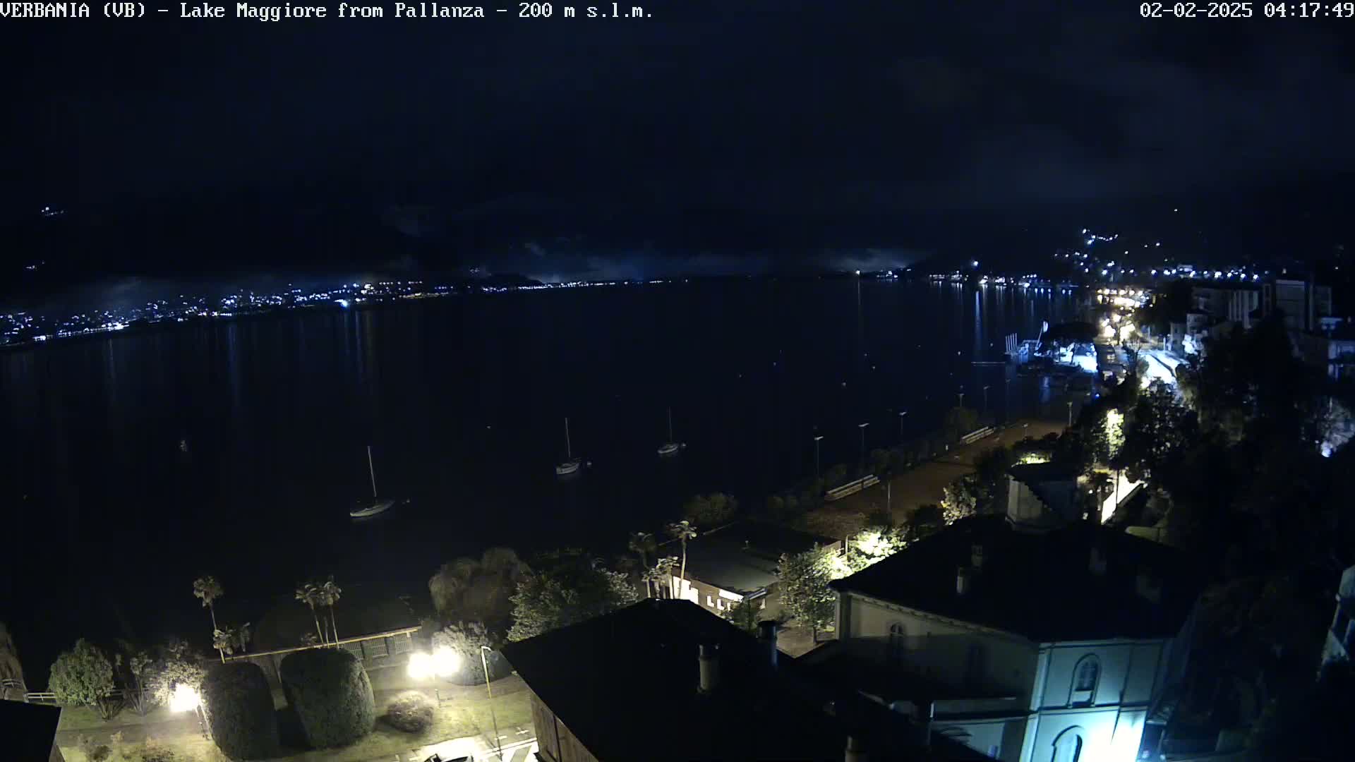 A nighttime view of a lake with lights on the opposite shore and a few boats on the water, with buildings and streetlights visible in the foreground under a partly cloudy sky.
