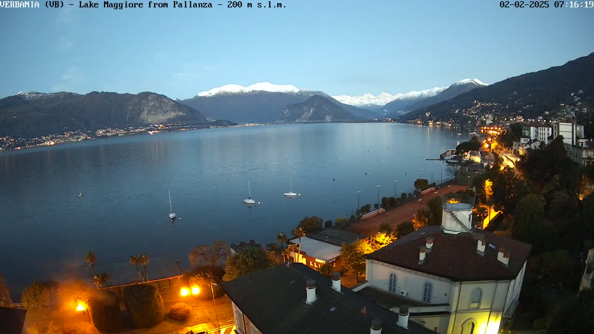 A twilight view of Lake Maggiore, with snow-capped mountains in the background and a town illuminated along the shoreline.
