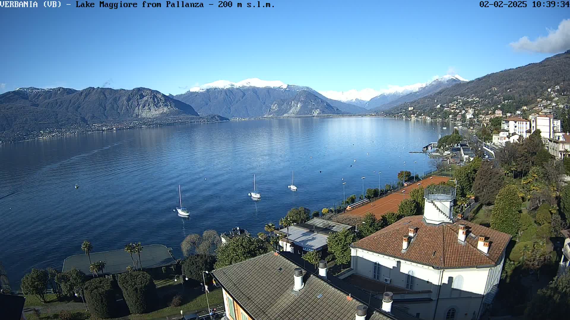A clear, sunny day overlooks a calm lake with snow-capped mountains in the background, several sailboats on the water, and a building with a red tile roof in the foreground.
