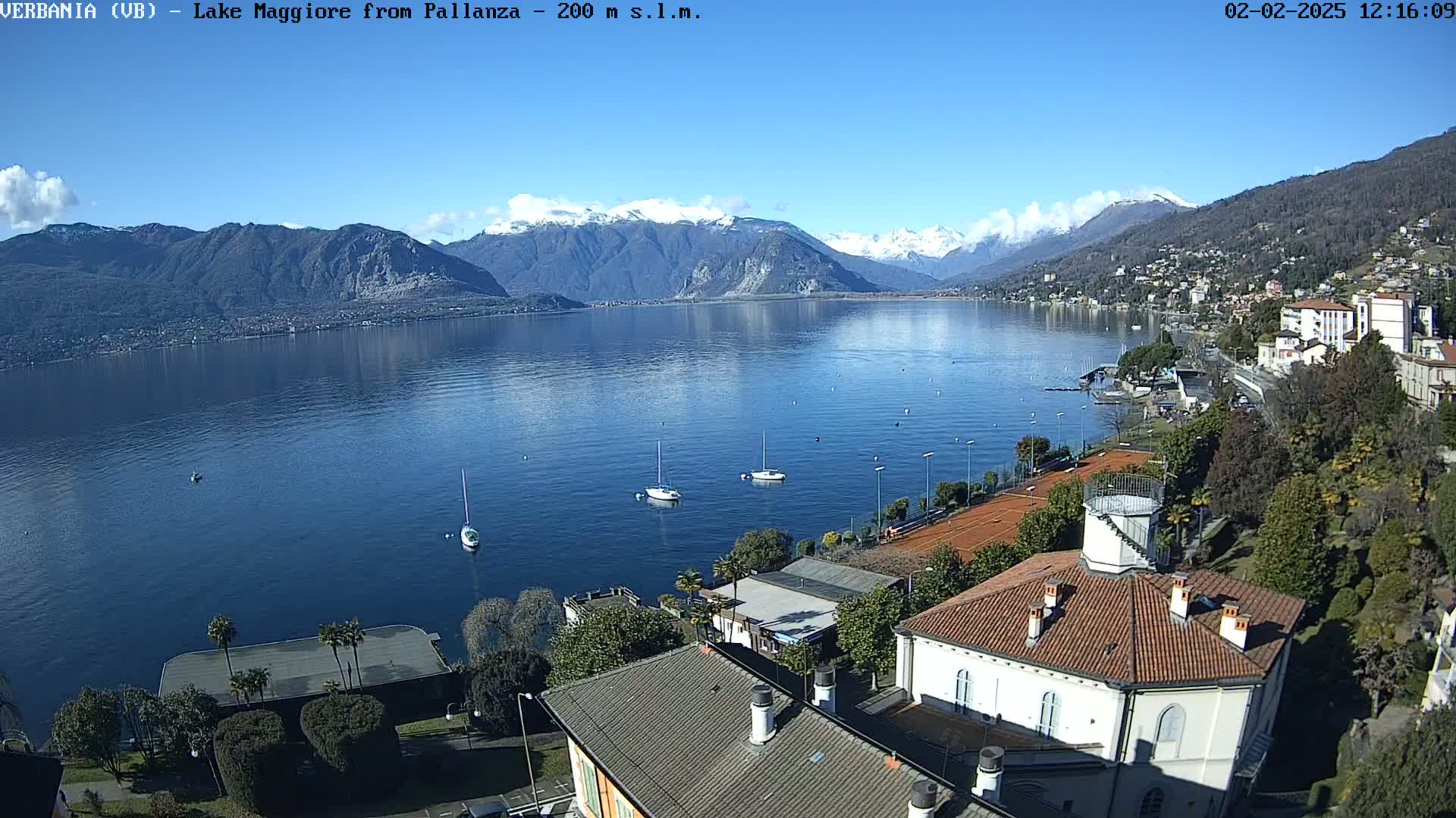 A sunny day overlooking a calm lake with snow-capped mountains in the background, showing sailboats on the water and buildings along the shoreline.