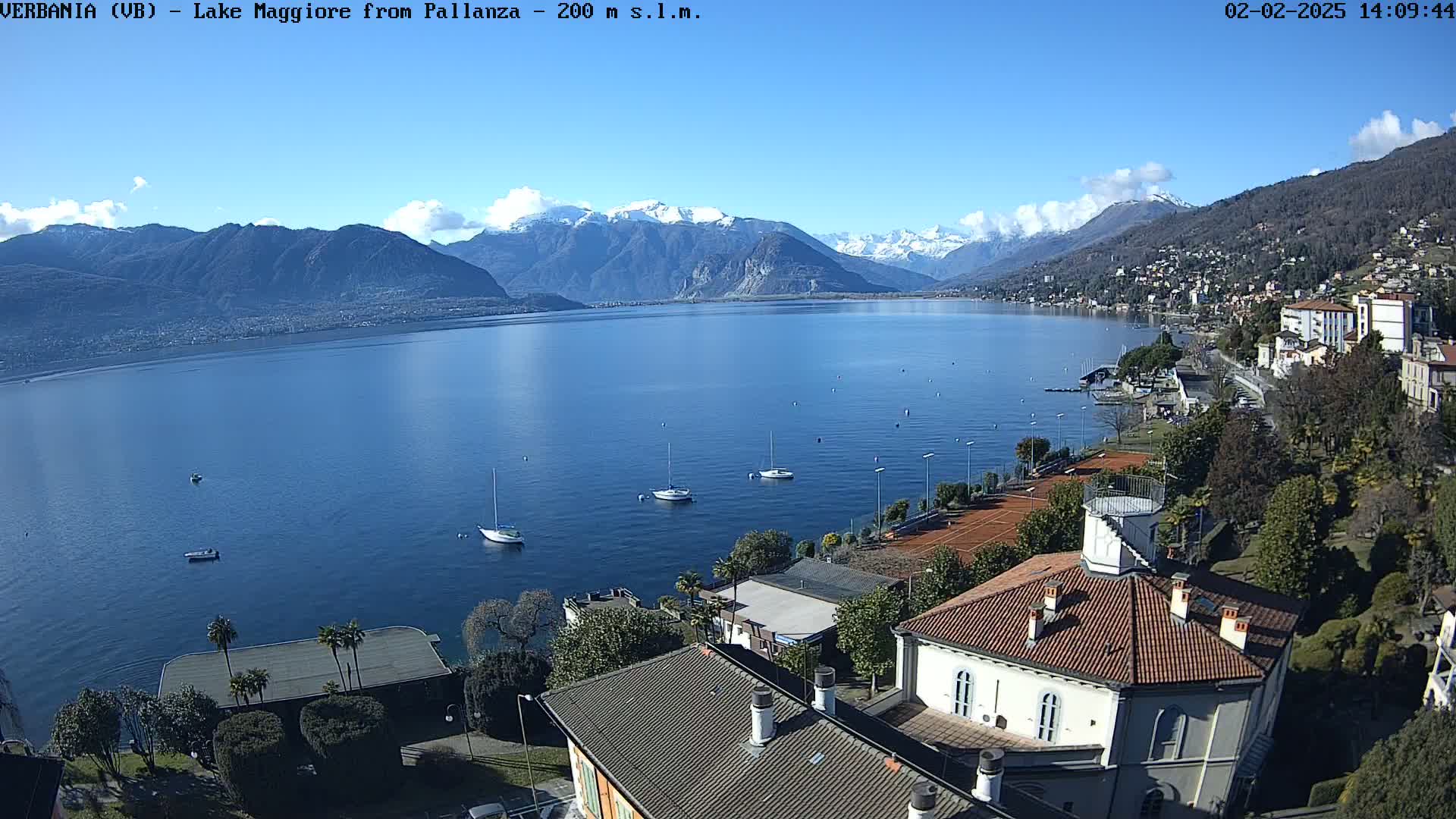 An aerial view shows a calm lake with sailboats, a town with tennis courts, and snow-capped mountains in the background under a clear blue sky.