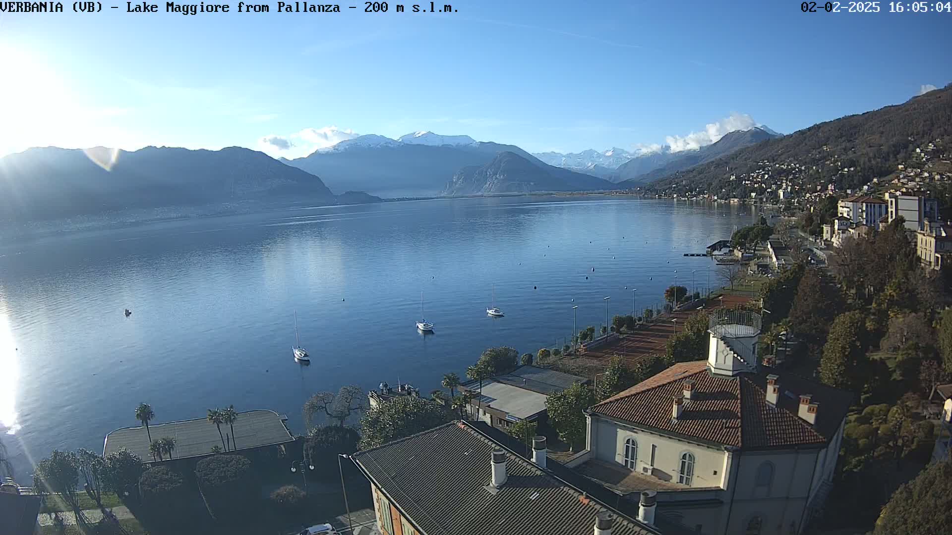 A clear, sunny day overlooking a calm lake with snow-capped mountains in the distance, featuring several boats and a town on the lakeshore.