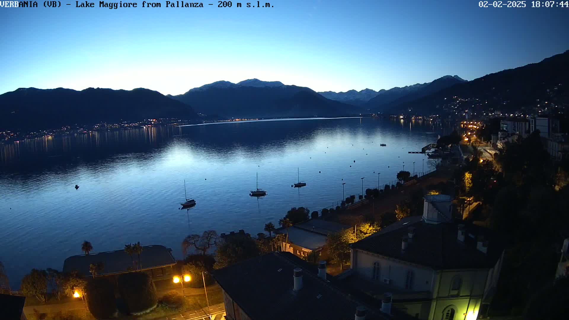A nighttime view of a calm lake with several boats, a town with lights on the shore, and mountains in the background under a clear twilight sky.