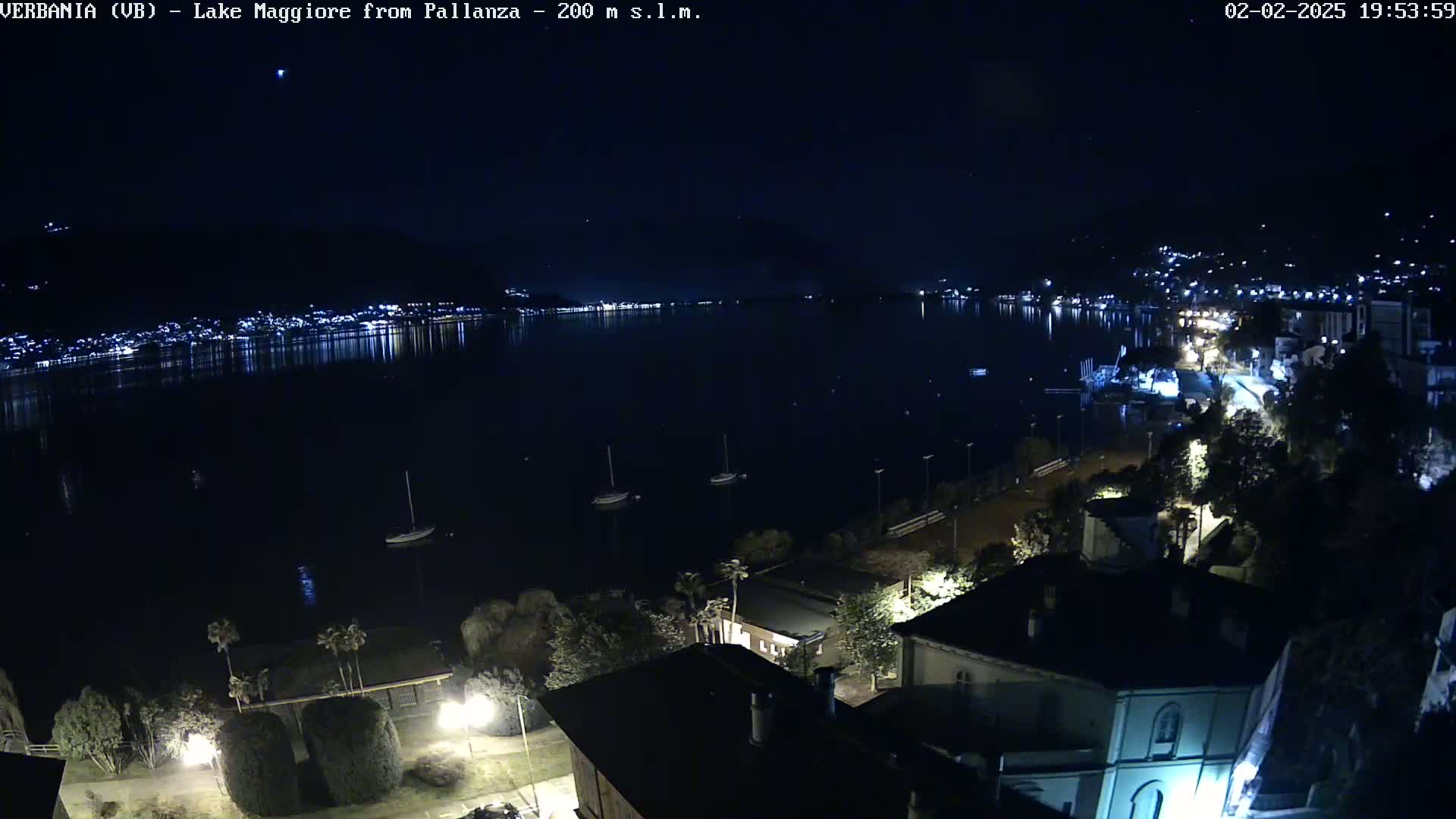 A nighttime view of a lake with a city and hills in the background, a few boats on the water, and buildings illuminated along the shoreline.