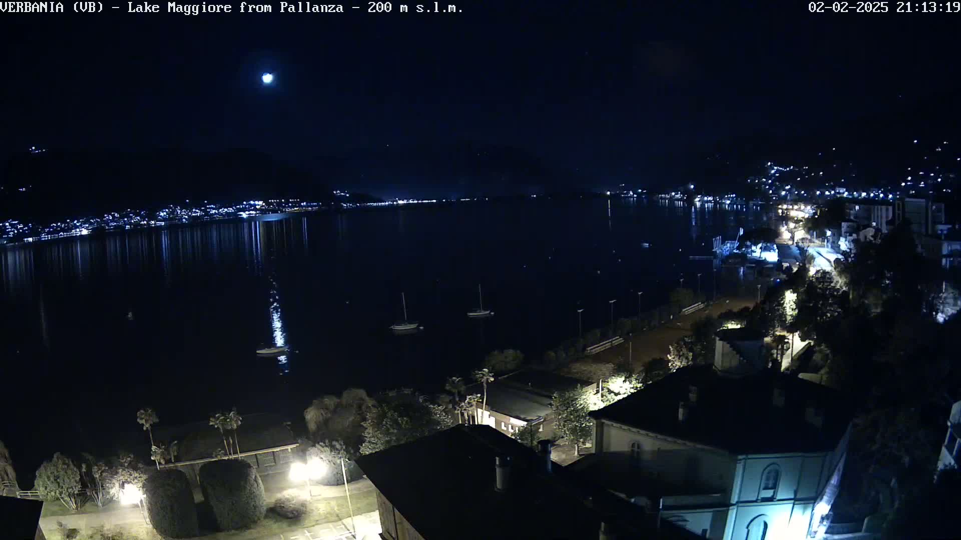 A nighttime view of a lake with a town's lights reflecting on the water under a clear, moonlit sky.