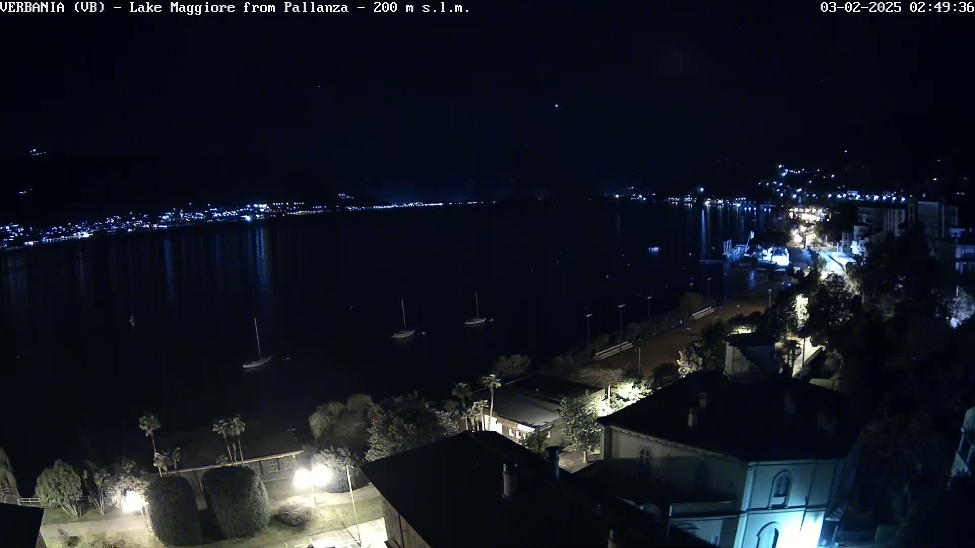 A nighttime view of a lake with a town’s lights visible across the water, several sailboats anchored in the foreground, and buildings and trees illuminated along the shore.