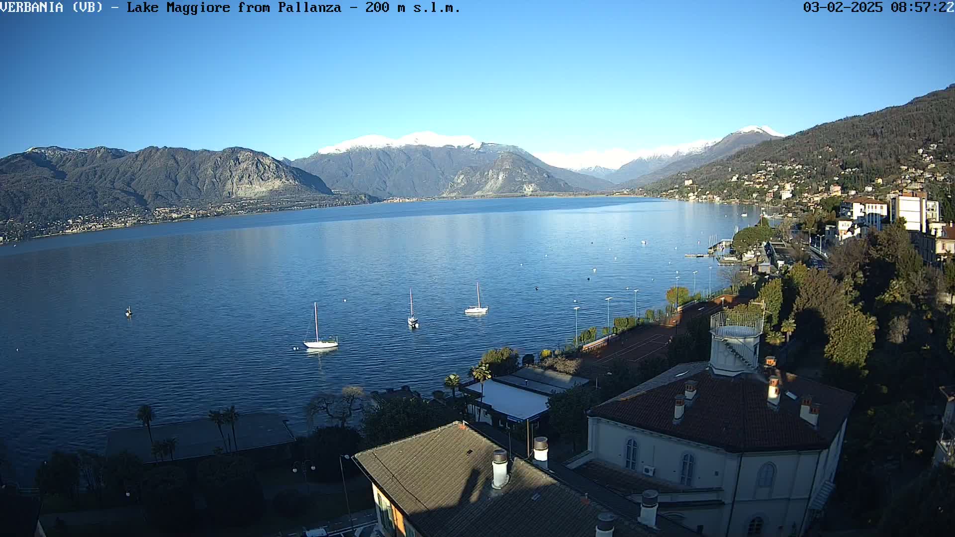 A clear, sunny day overlooking a large lake with snow-capped mountains in the background, several sailboats on the water, and a town with buildings visible along the shoreline.