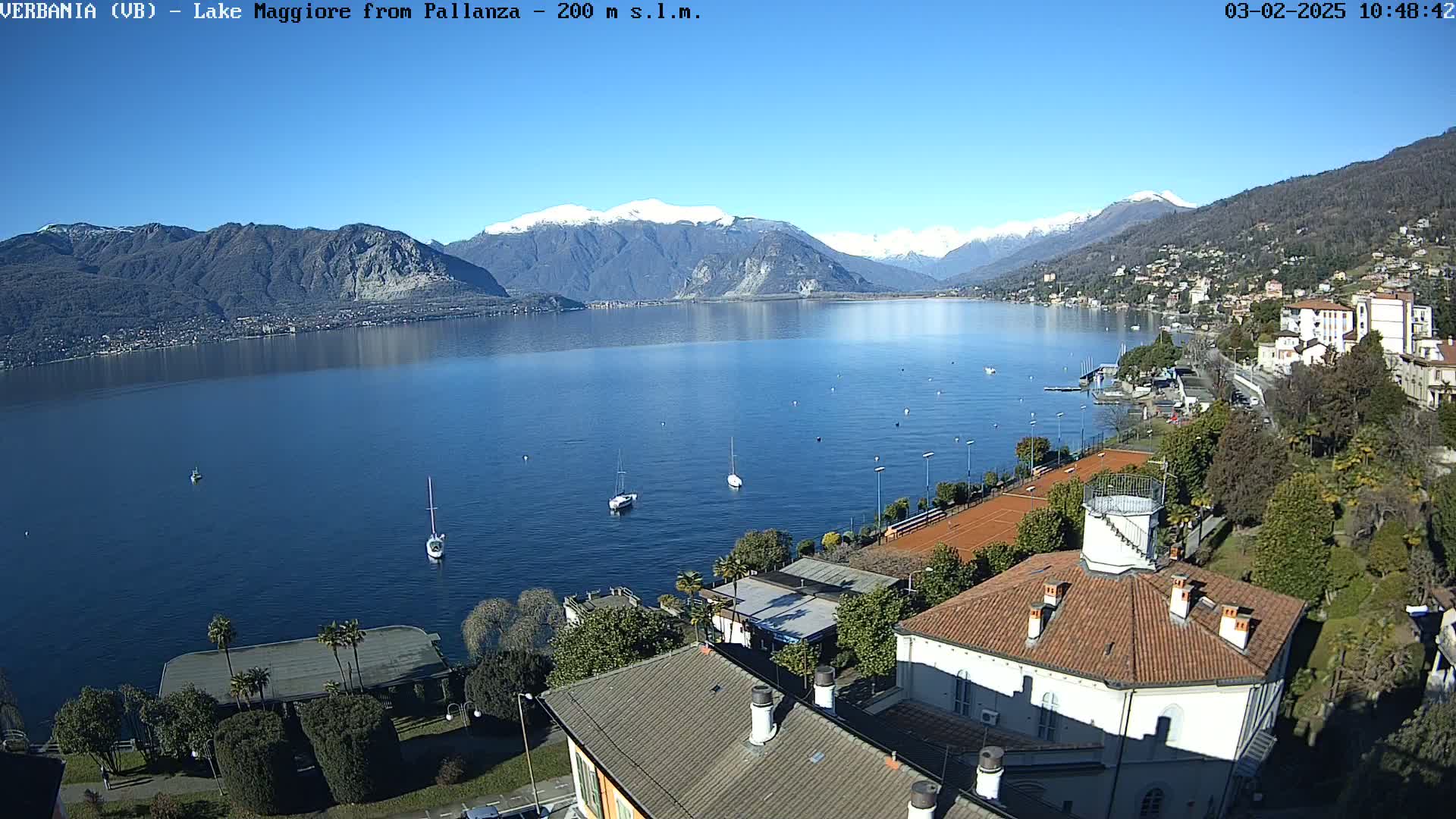 A sunny day overlooking a calm lake with snow-capped mountains in the distance, featuring a lakeside town with buildings, trees, and tennis courts.