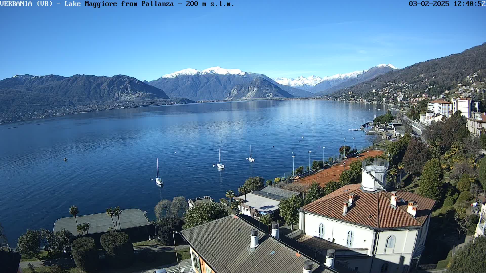 A clear, sunny day overlooks a calm lake with snow-capped mountains in the background, several sailboats on the water, and buildings and tennis courts along the shoreline.