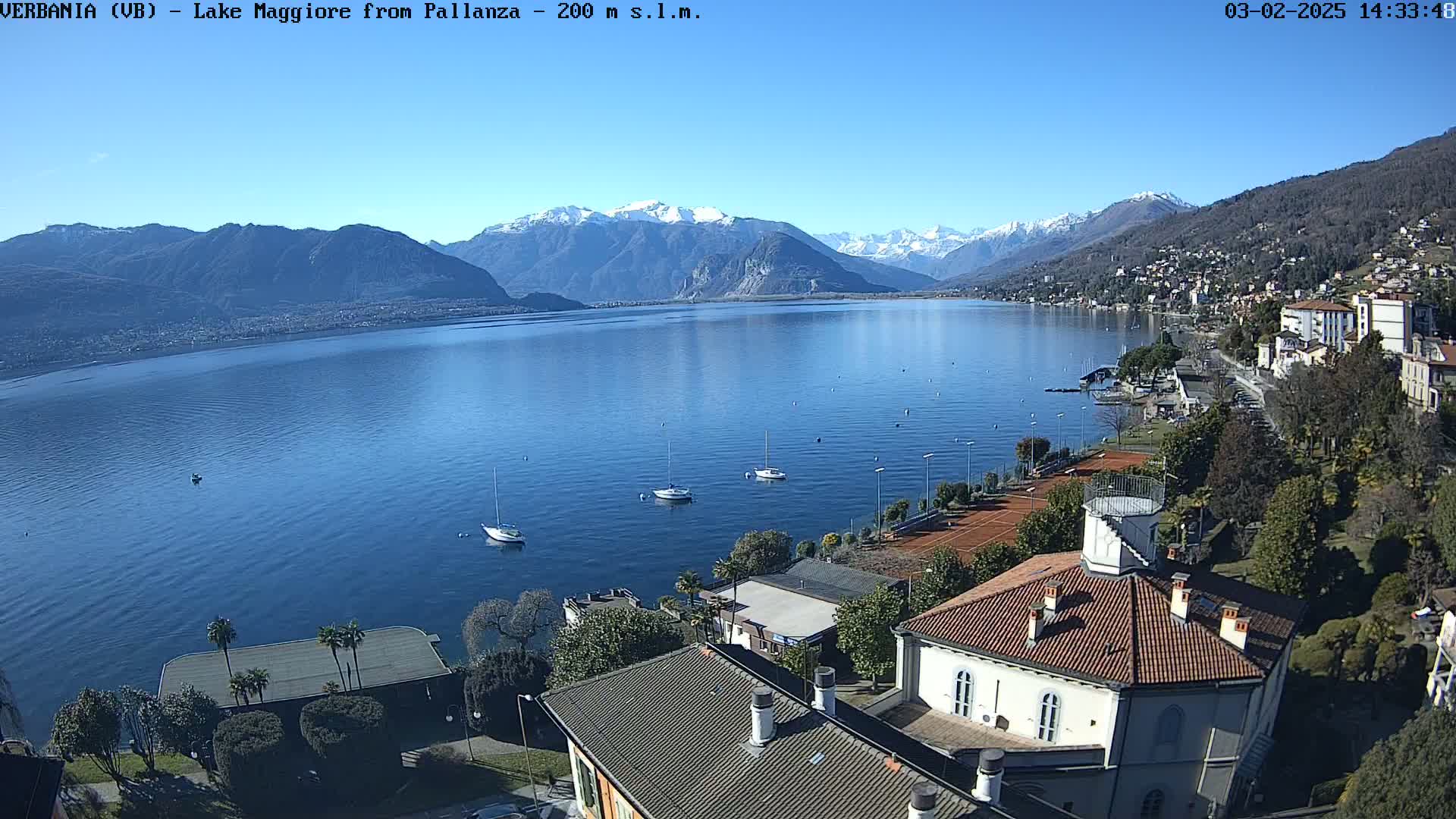 A sunny day overlooking a calm lake with snow-capped mountains in the distance, featuring several sailboats on the water and buildings along the shoreline.