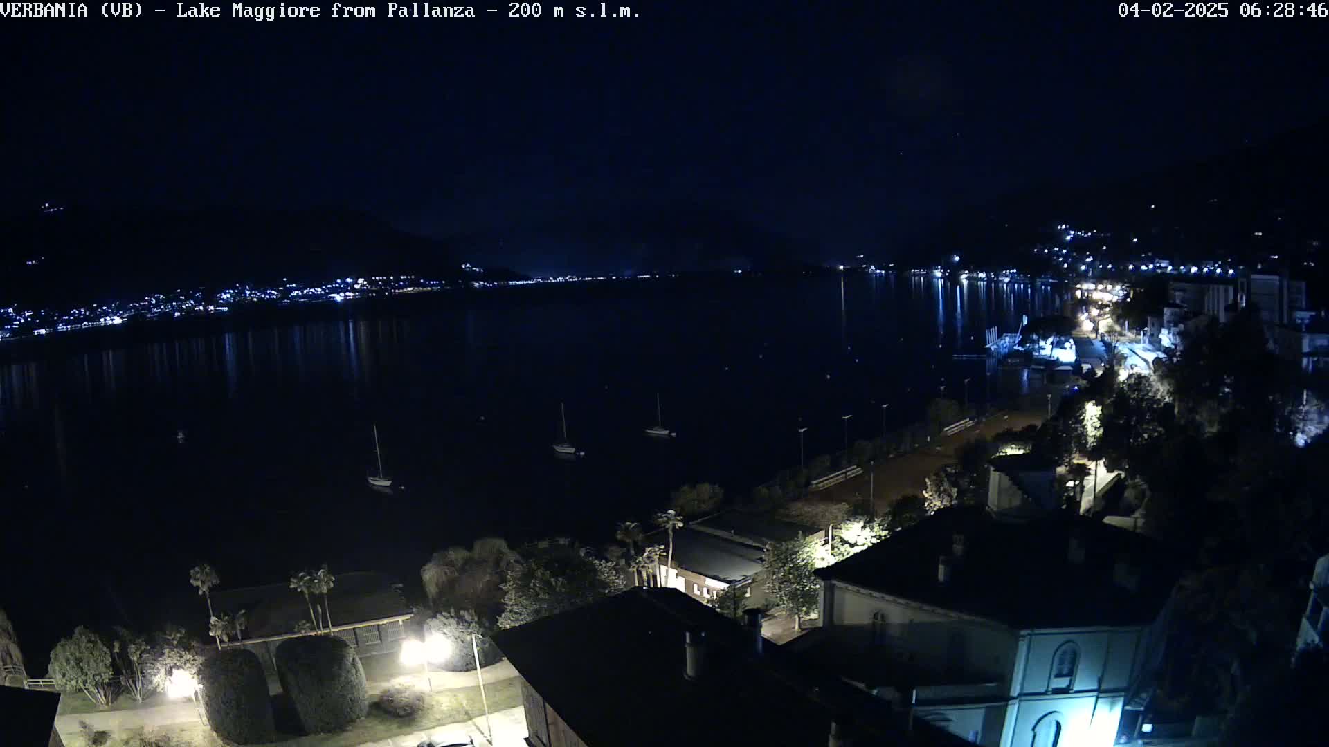 A nighttime, aerial view of a calm lake reflecting city lights, with boats moored nearby and buildings illuminated along the shore.