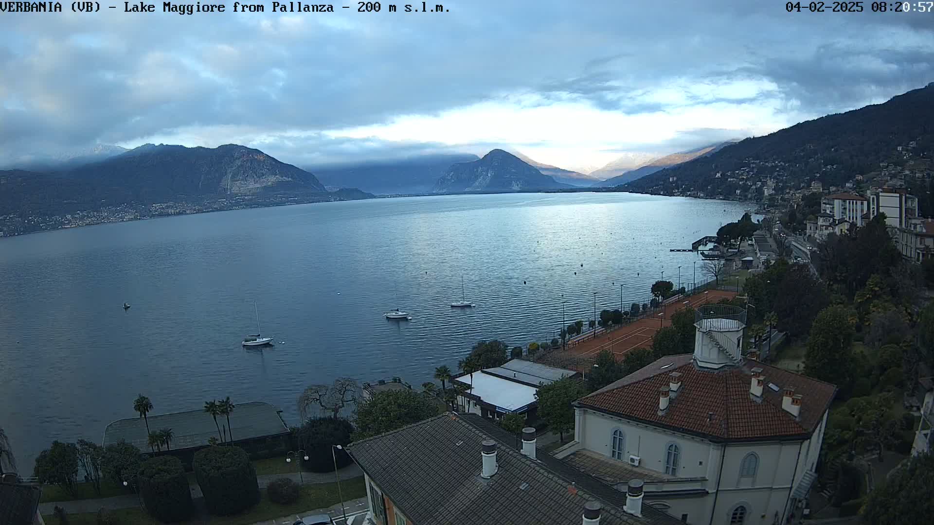 A partly cloudy day overlooking a calm lake with several small boats, mountains in the distance, and a town along the shoreline, including tennis courts and buildings.