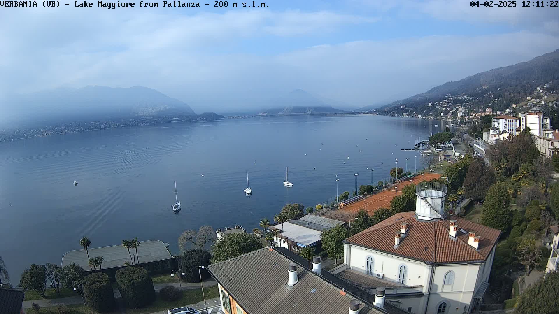 A tranquil lake scene under a mostly clear sky shows several sailboats on the water, a shoreline with buildings and tennis courts, and mountains in the distance.
