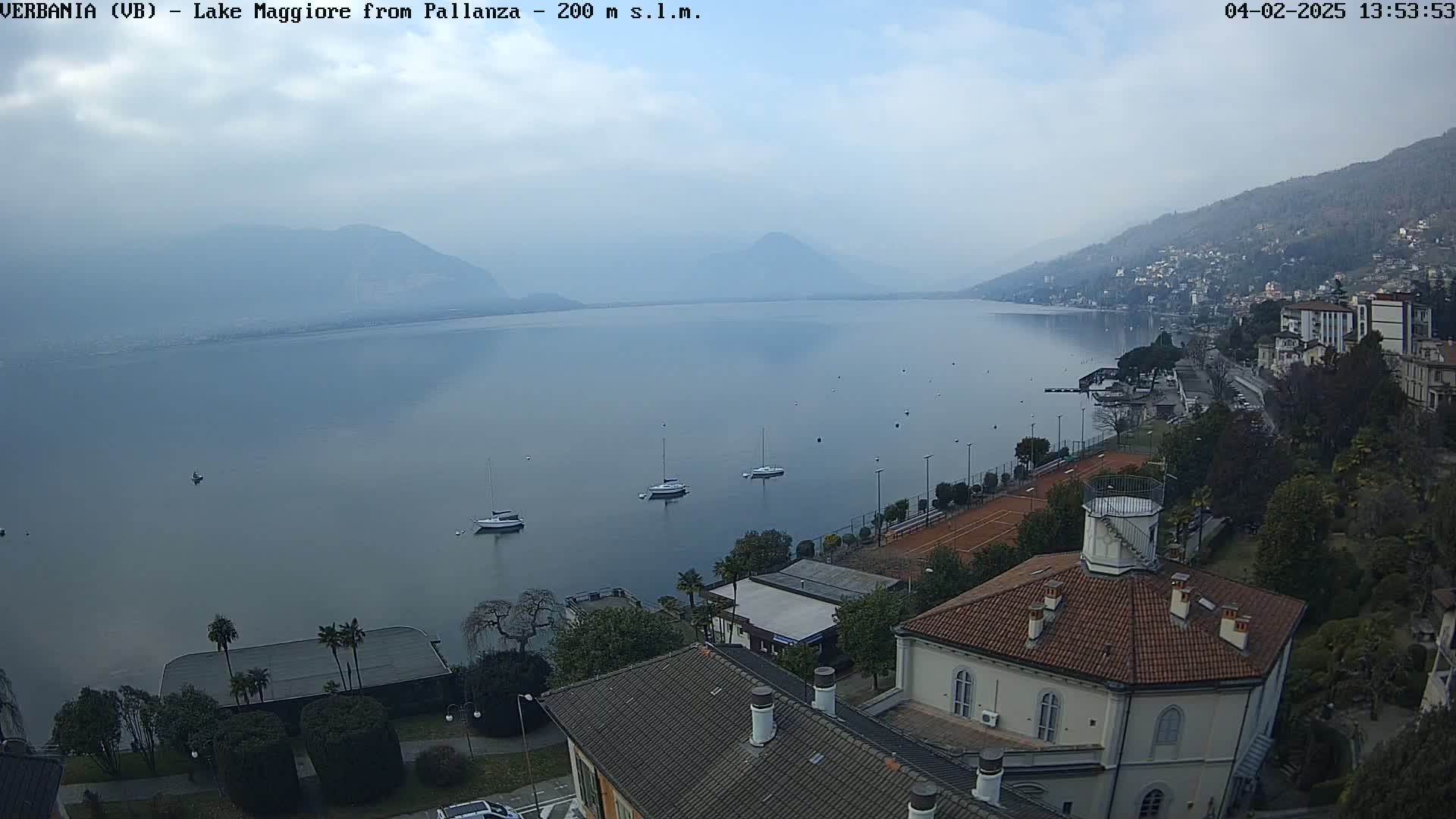 A hazy, overcast day overlooks a calm lake with several sailboats and a town nestled along the shoreline, featuring buildings, tennis courts, and lush greenery.
