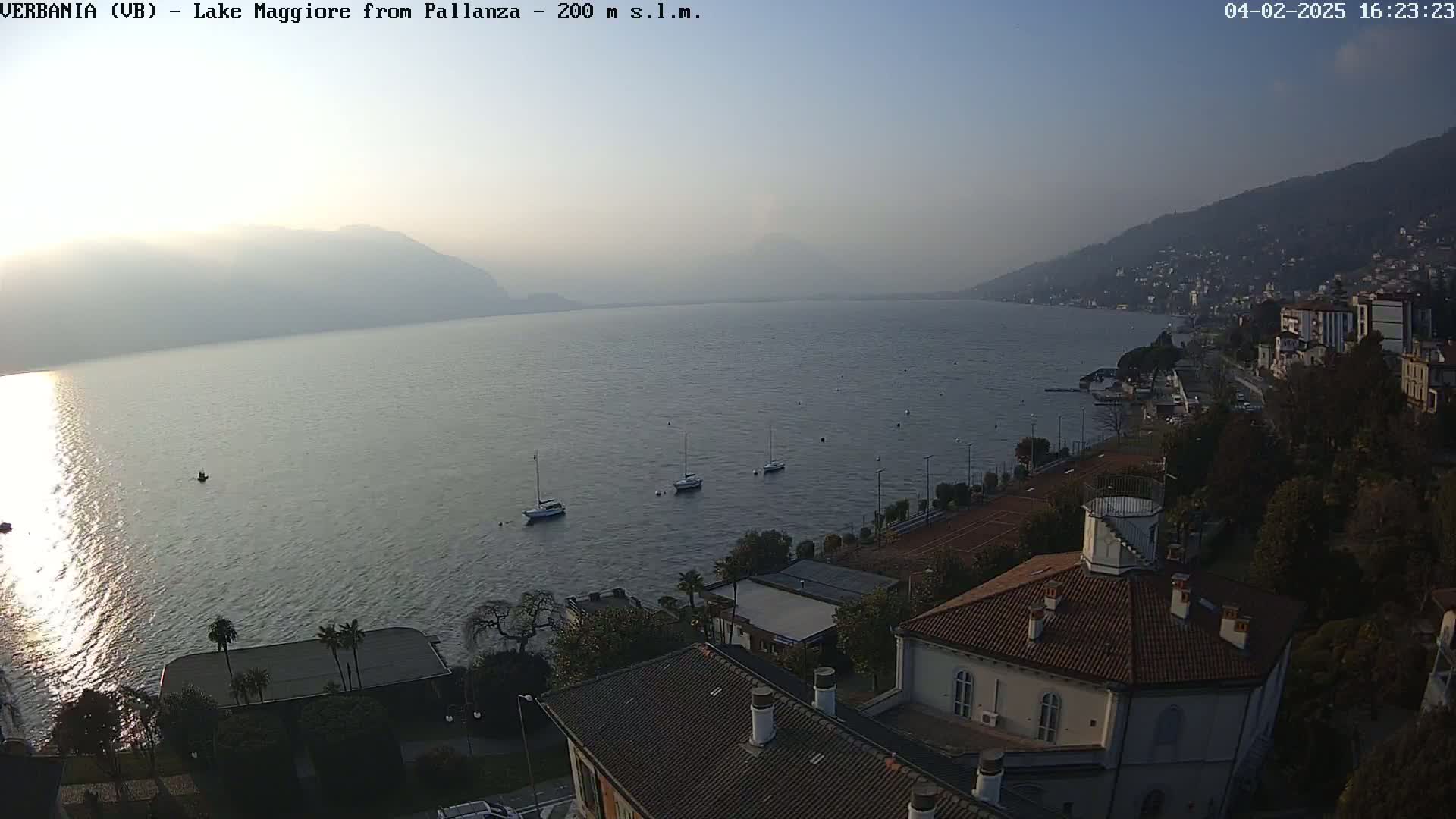 A hazy, calm lake stretches to distant mountains under a pale sky, with a few sailboats on the water and buildings lining the shore.