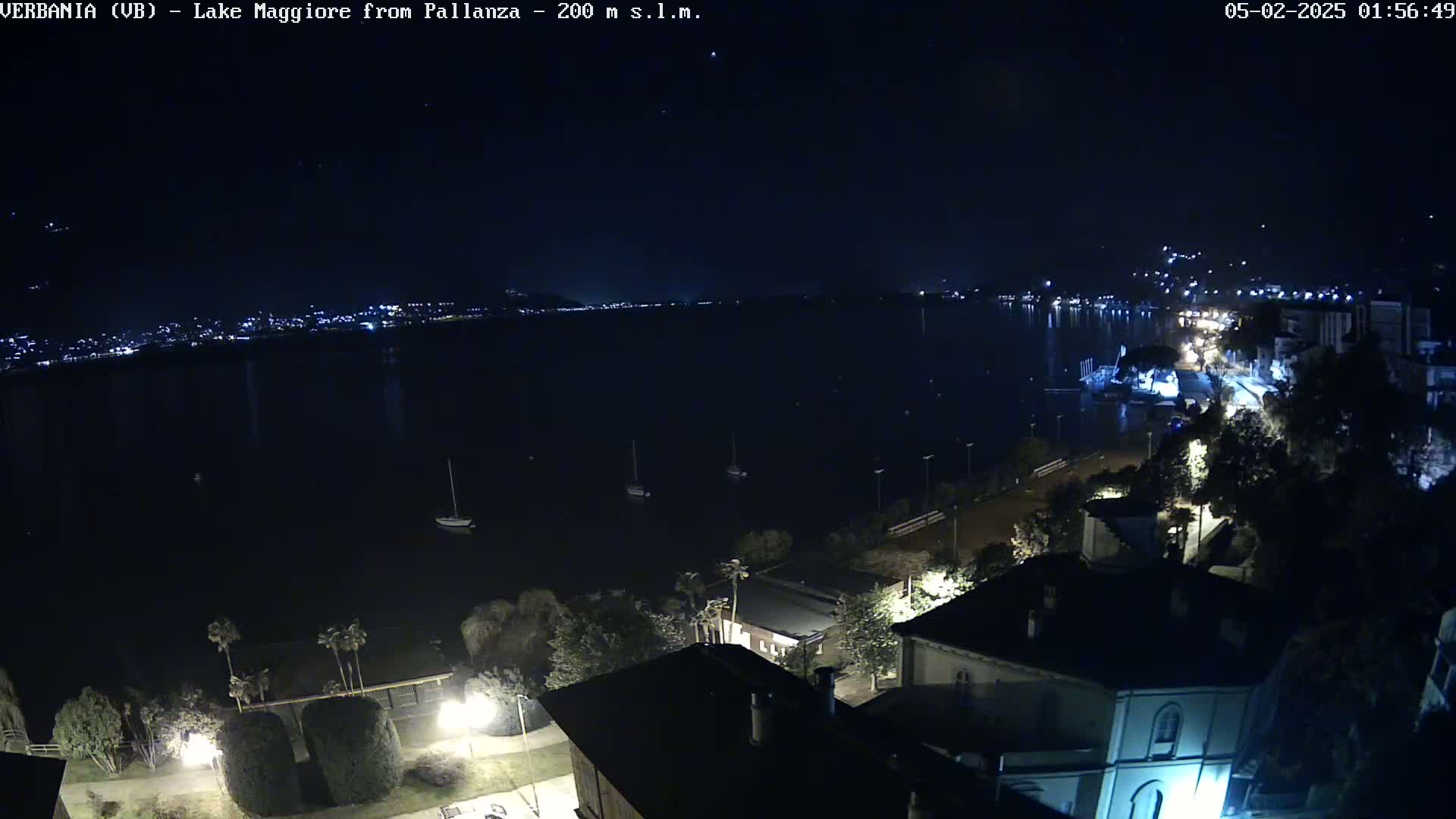 A nighttime view of a dark lake with a few boats and a shoreline with buildings and streetlights under a clear sky.