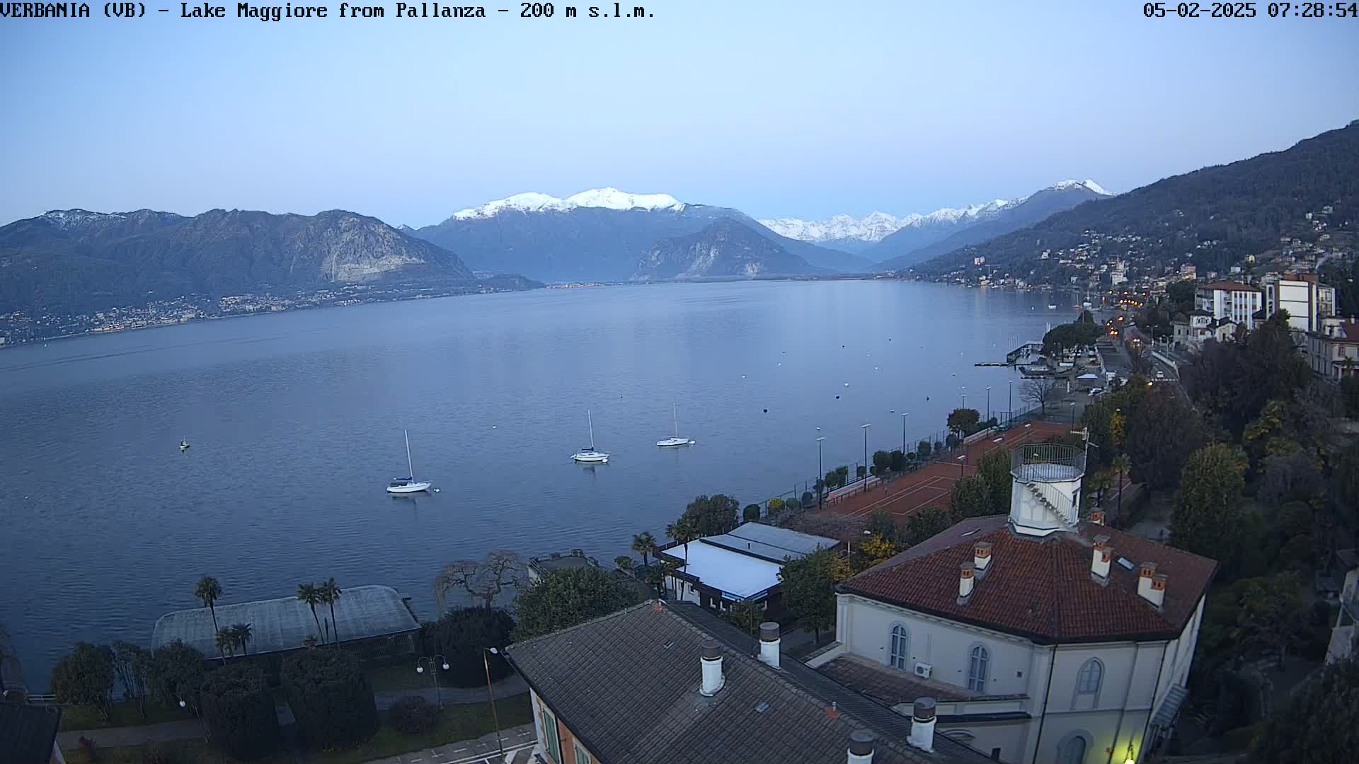 A calm lake stretches before snow-capped mountains under a twilight sky, with a few sailboats on the water and buildings along the shoreline.