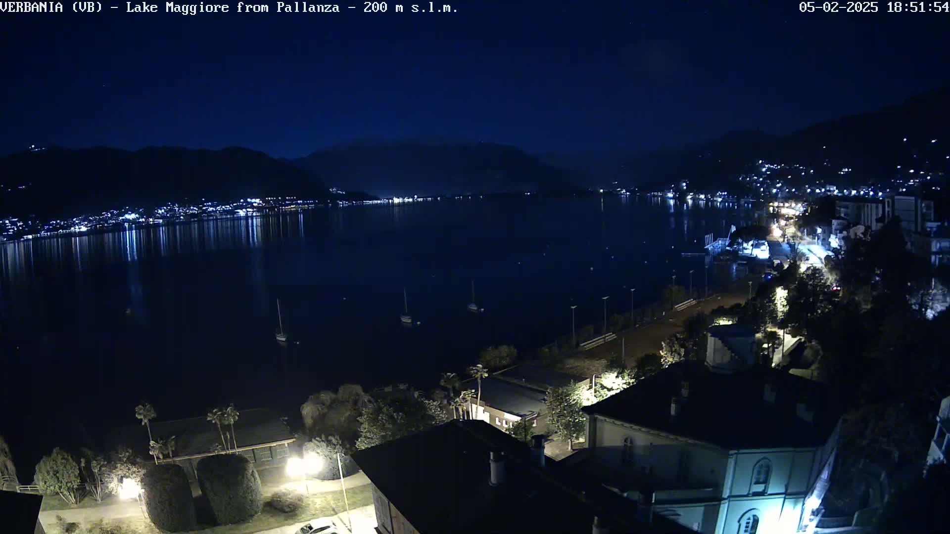 A nighttime view of a calm lake reflecting the lights of a town on its shore, under a clear, dark sky.