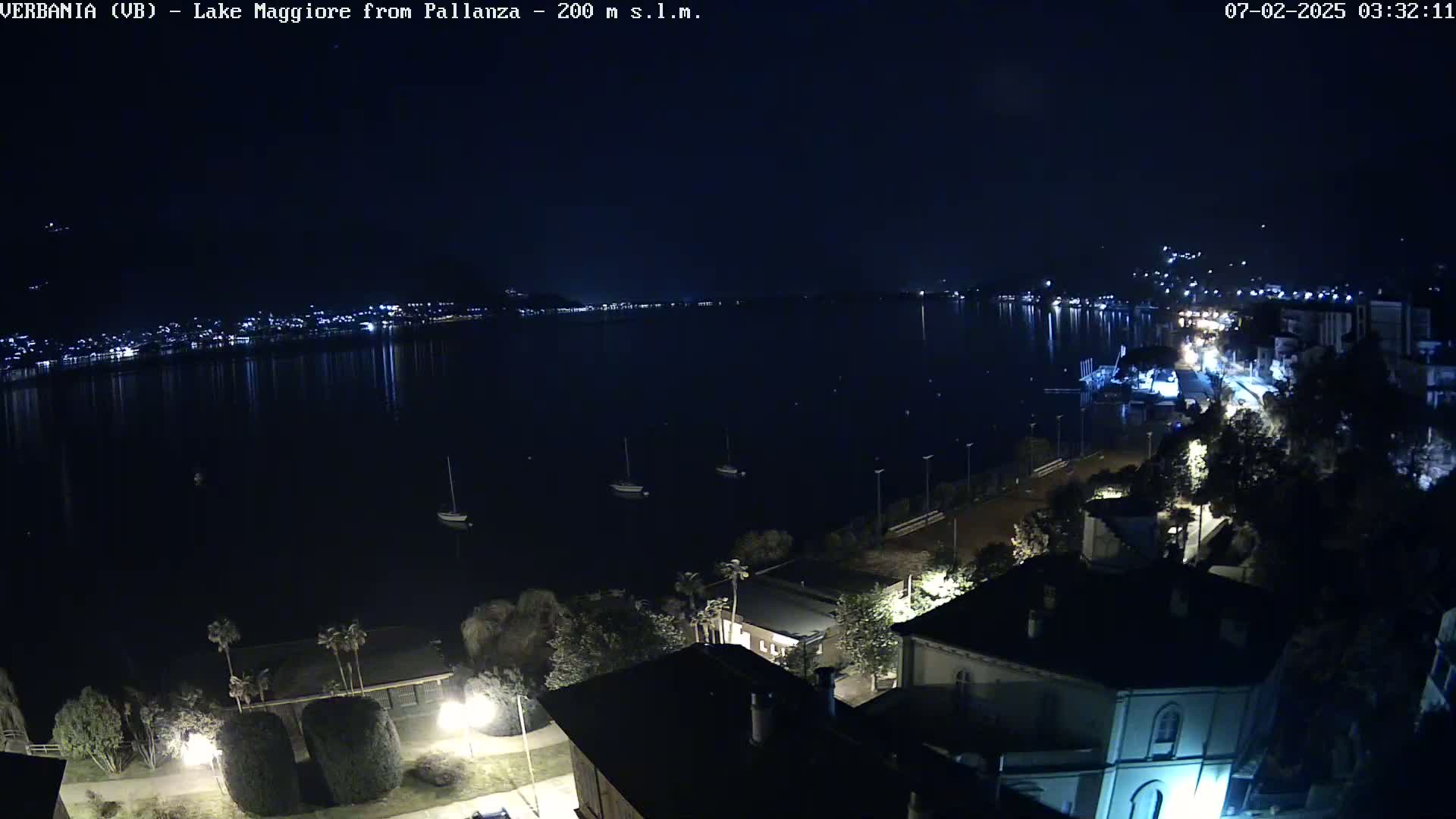 A nighttime, long shot of a lake with a town's lights reflecting on the water, a few boats on the lake, and buildings and trees on the shore.